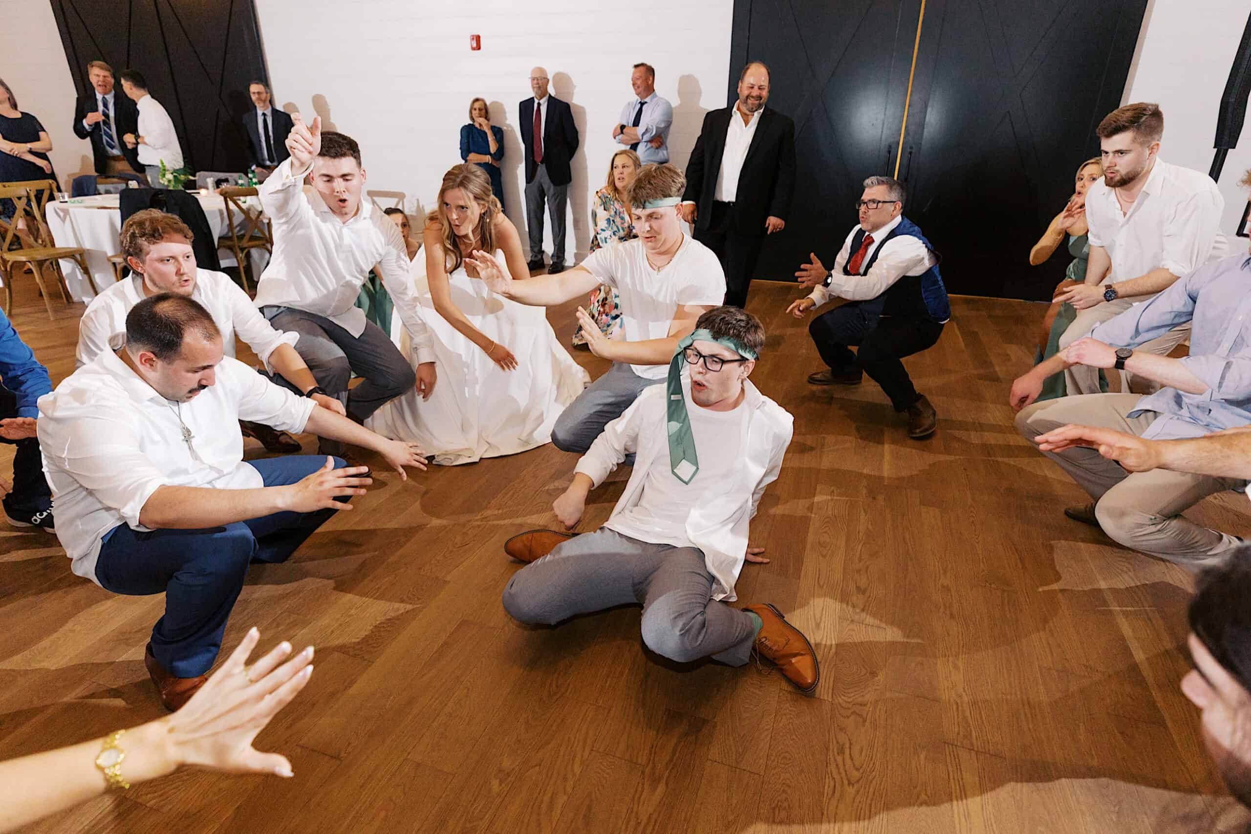 A group of people, some in dress shirts and ties, energetically dancing and posing on a wooden floor at Westwynd Gardens Spring Wedding, with onlookers in the background.