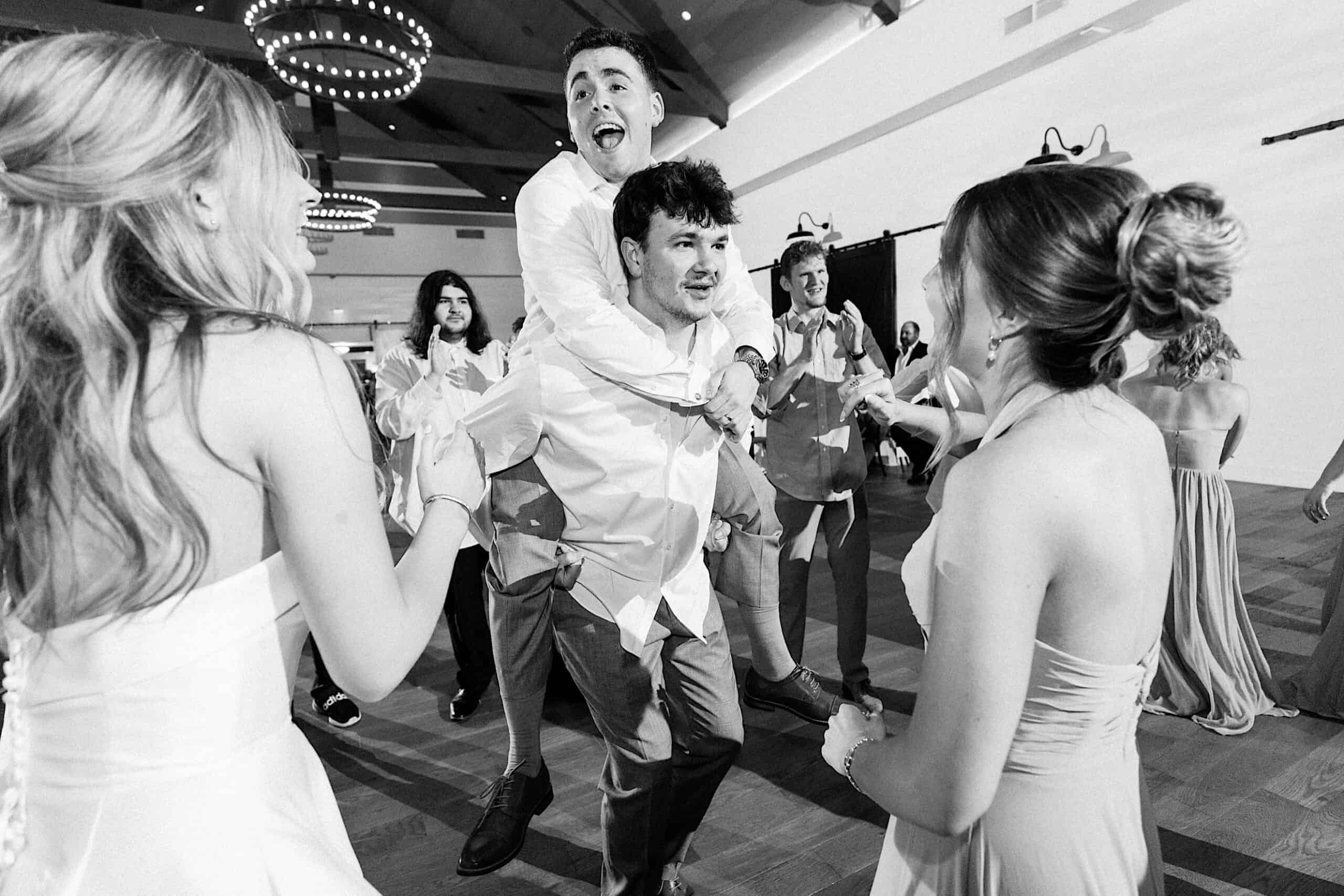 At a Westwynd Gardens Spring Wedding, guests dressed formally dance together; one man gives another a piggyback ride while others smile and clap around them.