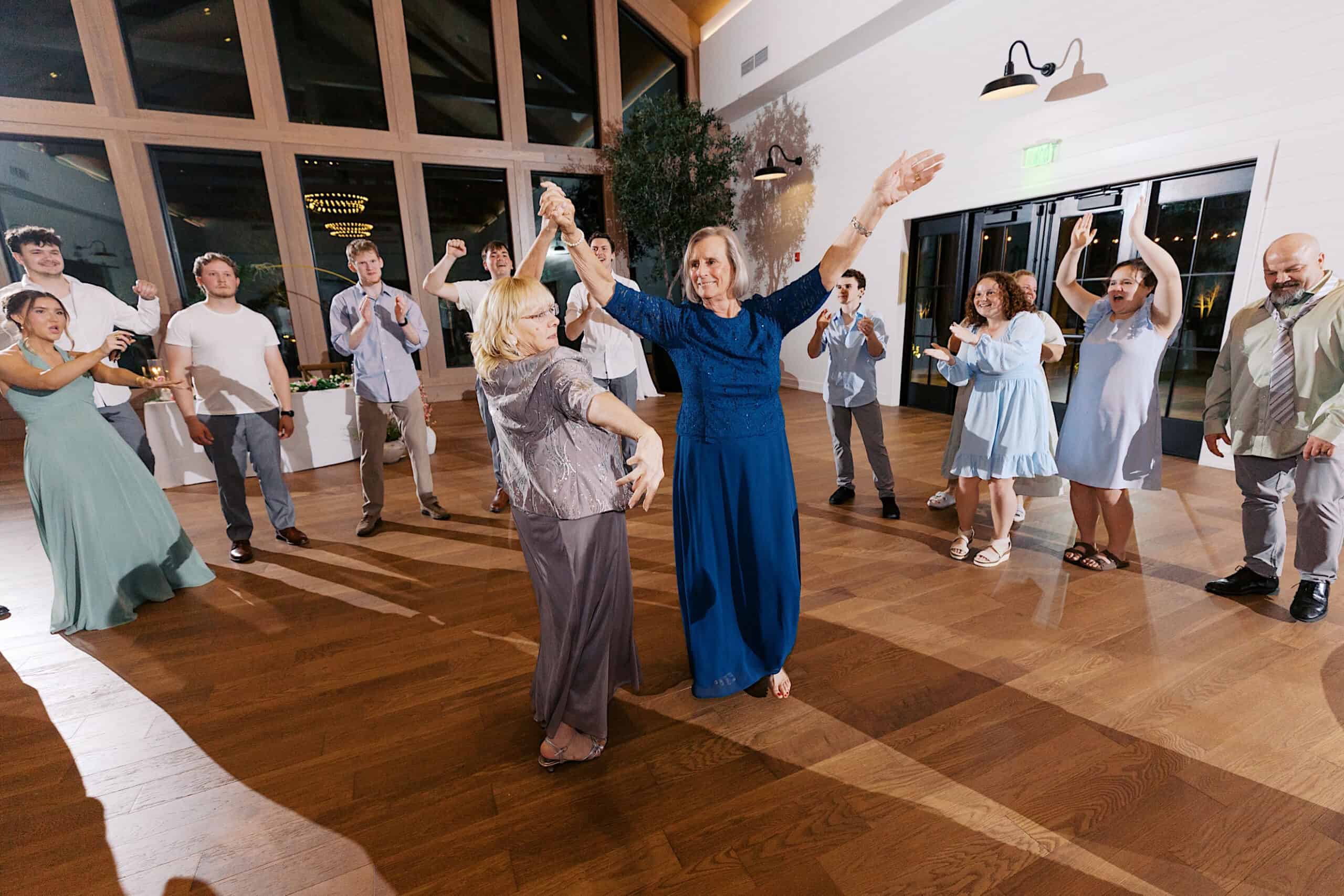 Two women dance in the center of a group at a Westwynd Gardens Spring Wedding, as guests form a circle around them, clapping and watching in a bright, spacious room with wooden floors.