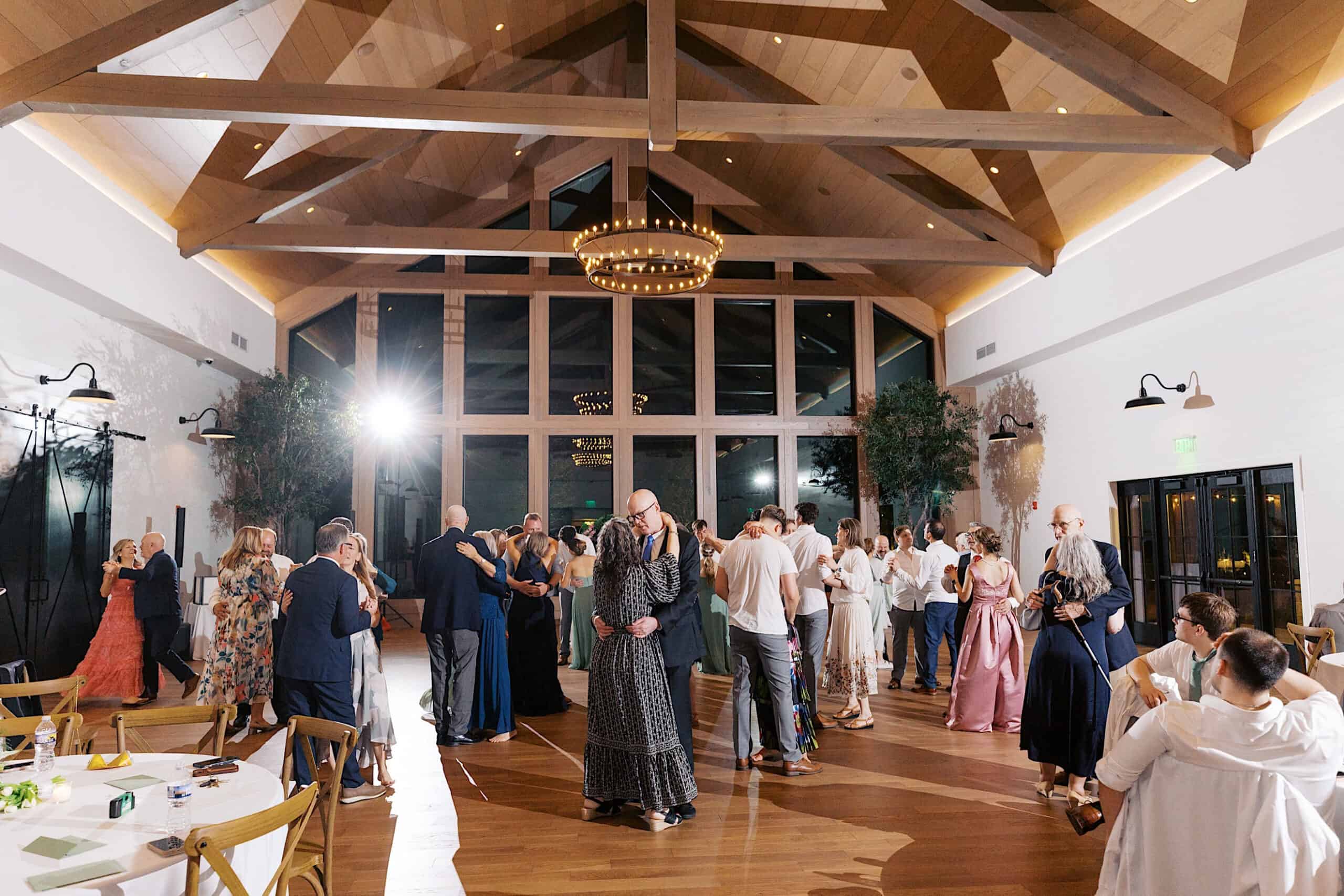 People are dancing and socializing in a spacious, well-lit venue with wooden beams, large windows, and elegant décor during a Westwynd Gardens Spring Wedding event.