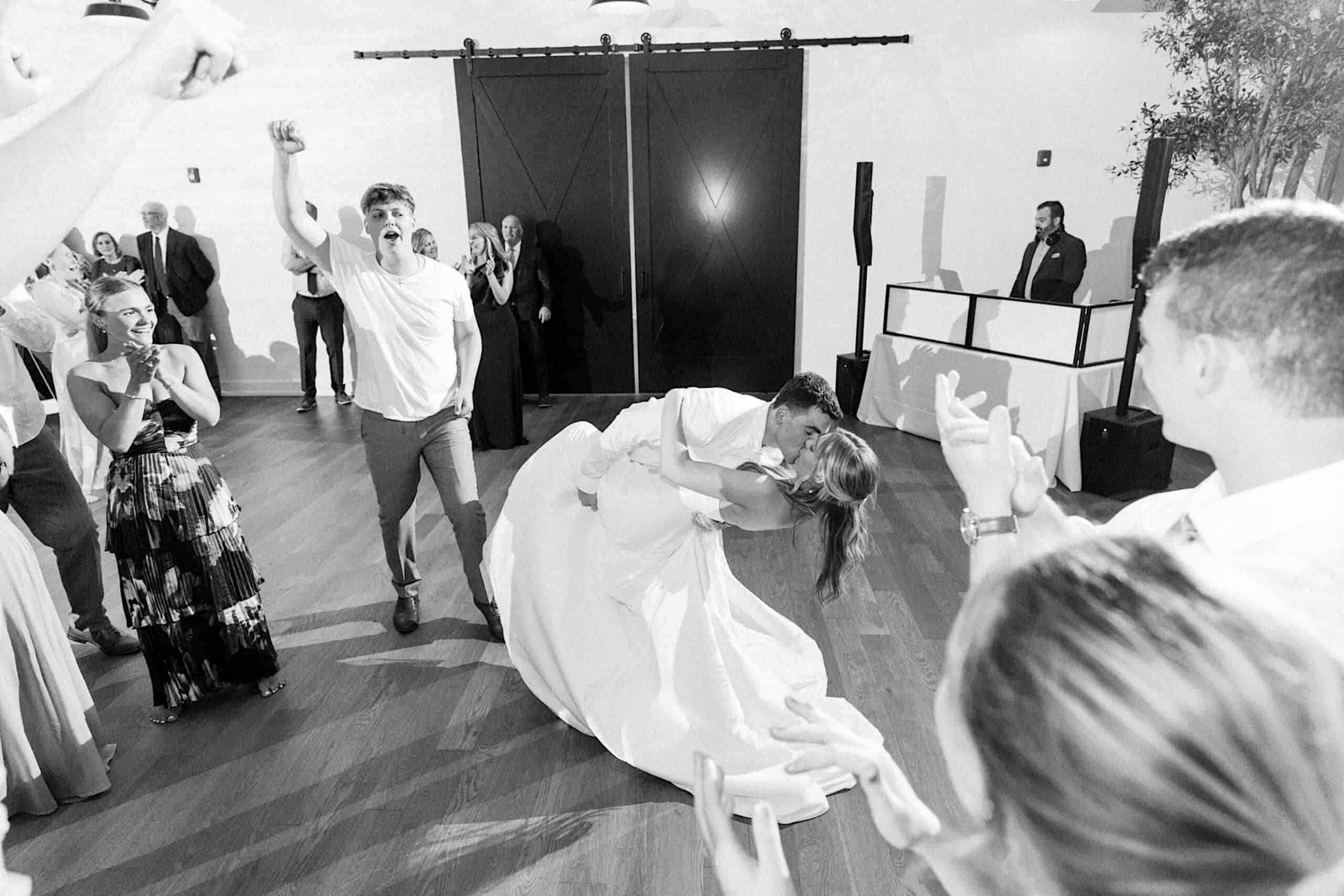 A couple in wedding attire shares a kiss while dancing at their Westwynd Gardens Spring Wedding, surrounded by cheering guests on a wooden floor in a well-lit reception hall.