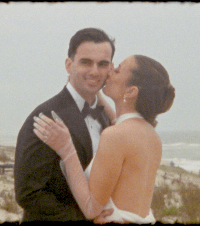 A woman in a white dress kisses a man in a black tuxedo on the cheek while standing on a beach with waves and sand dunes in the background, capturing an intimate moment from their Parker's Garage Wedding.