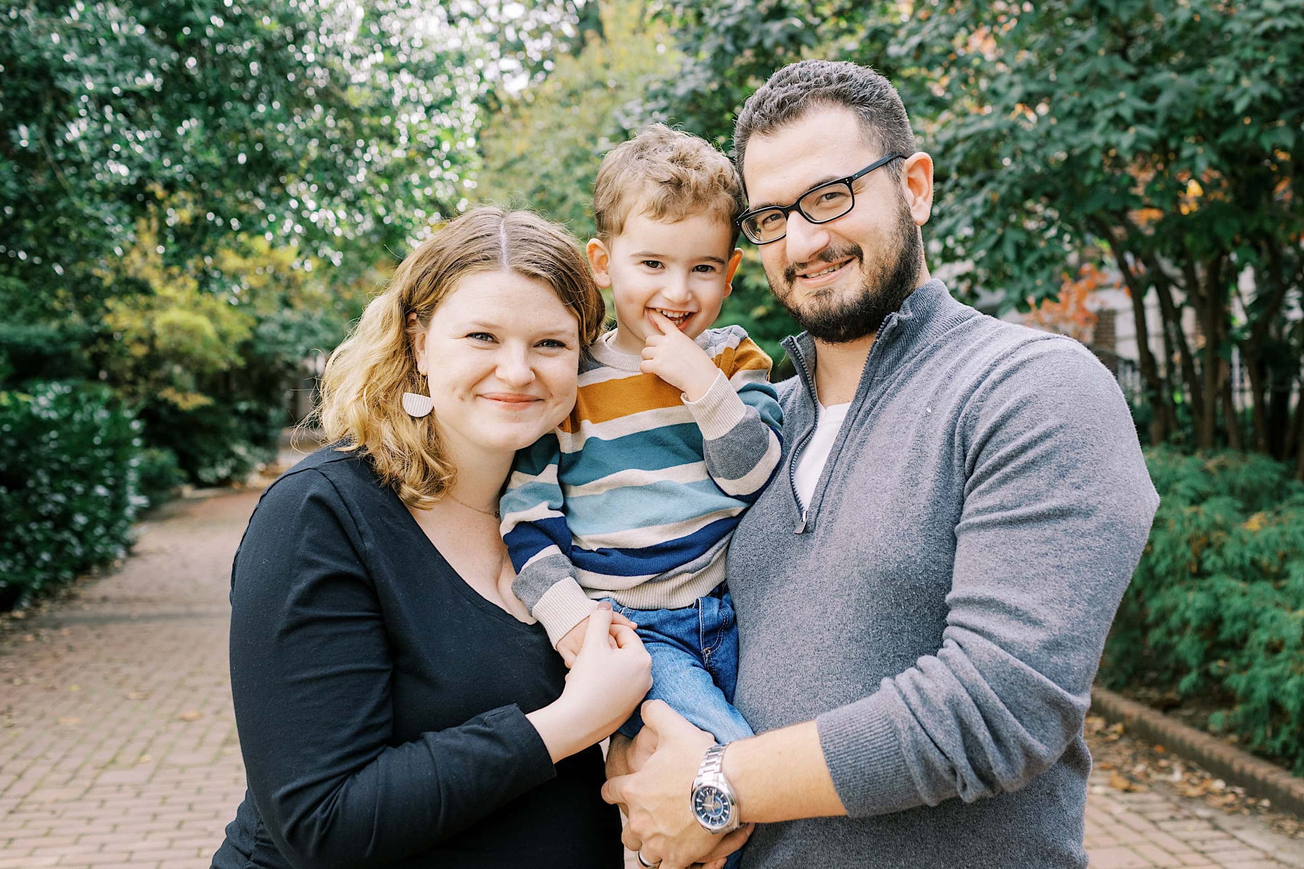 Three people, two adults and a child, stand close together outdoors on a brick path, smiling at the camera with green trees in the background—a classic moment captured in these Society Hill Family Photos.