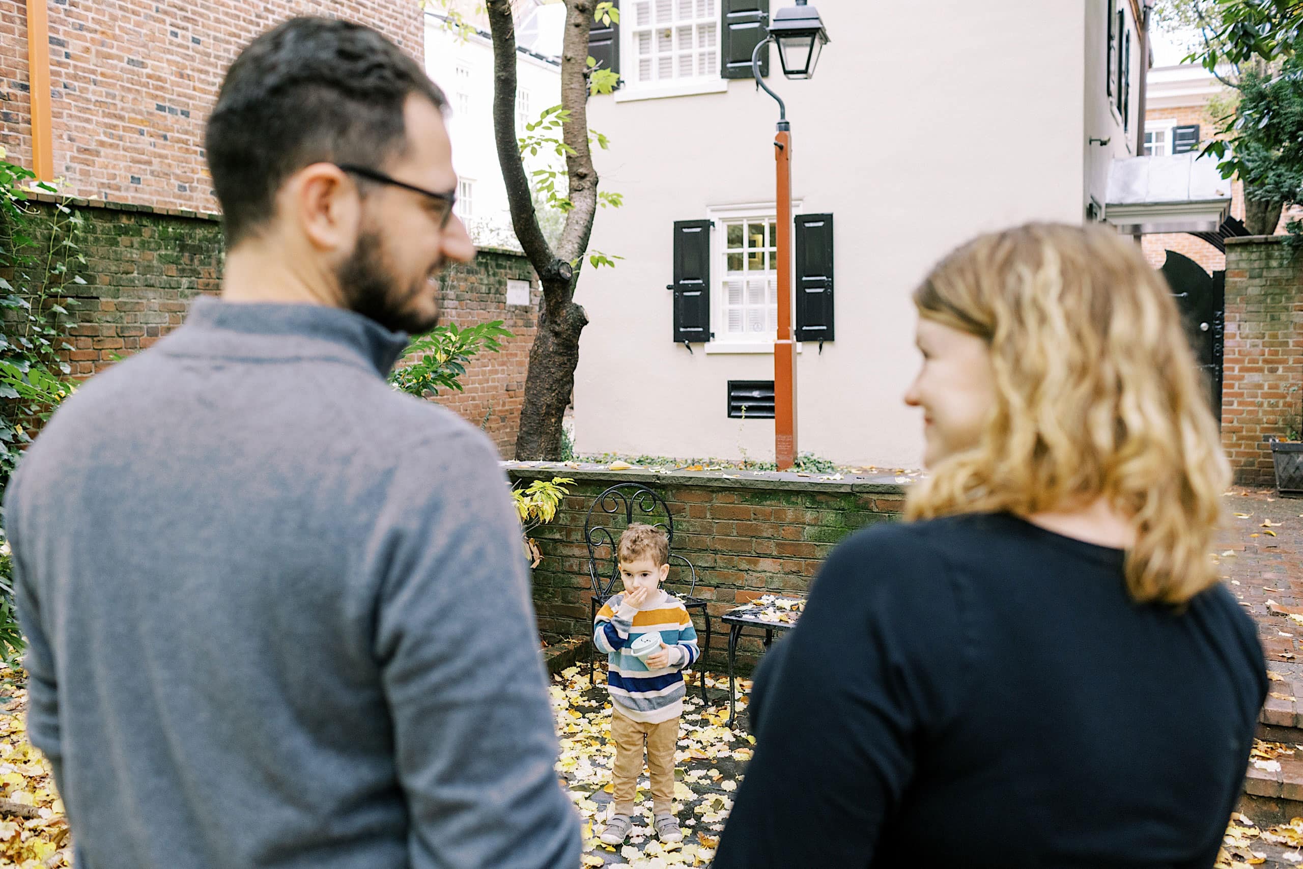 A child stands among fallen leaves in a courtyard while two adults, seen from behind, look toward him—a timeless moment beautifully captured in these Society Hill Family Photos.