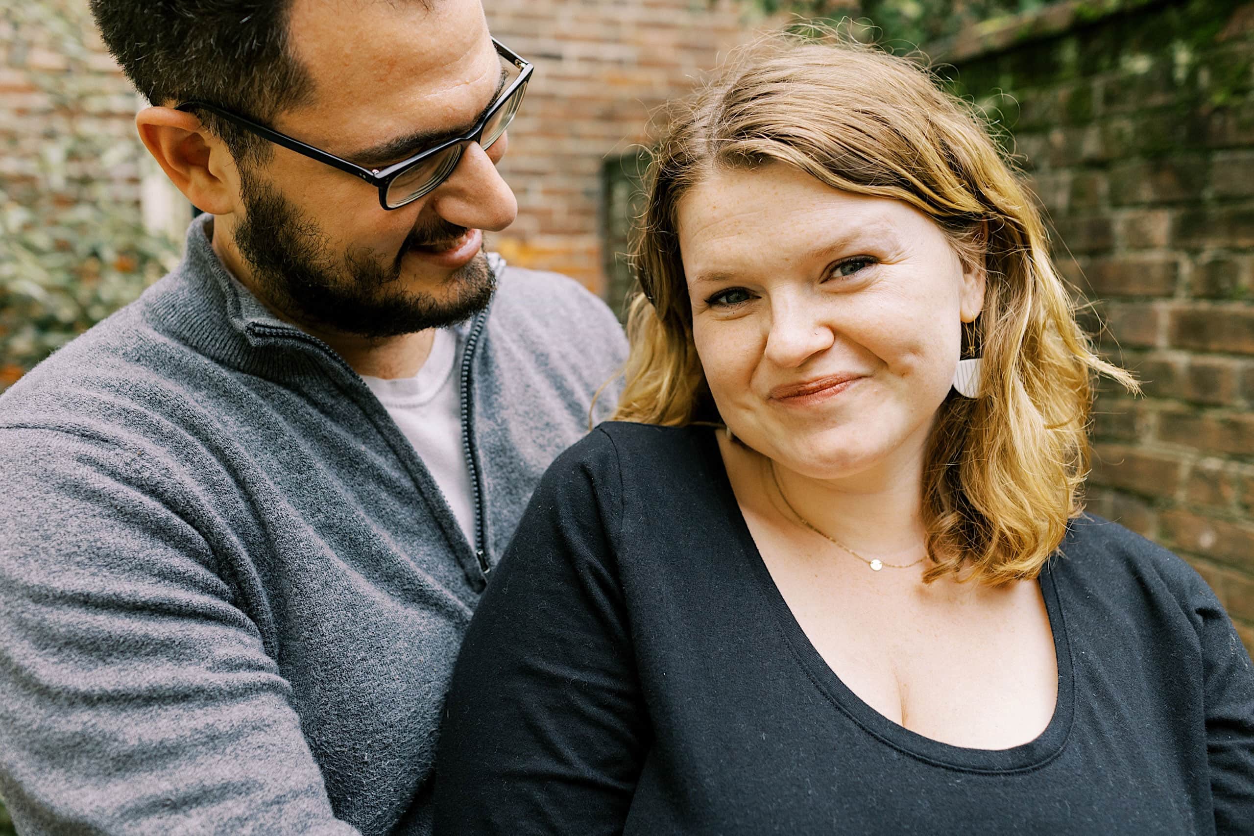 A man stands close behind a woman outdoors in what appears to be a relaxed moment perfect for Society Hill Family Photos. He is looking at her and smiling, while she faces the camera with a slight smile. Both wear casual clothing, and there is a brick wall in the background.