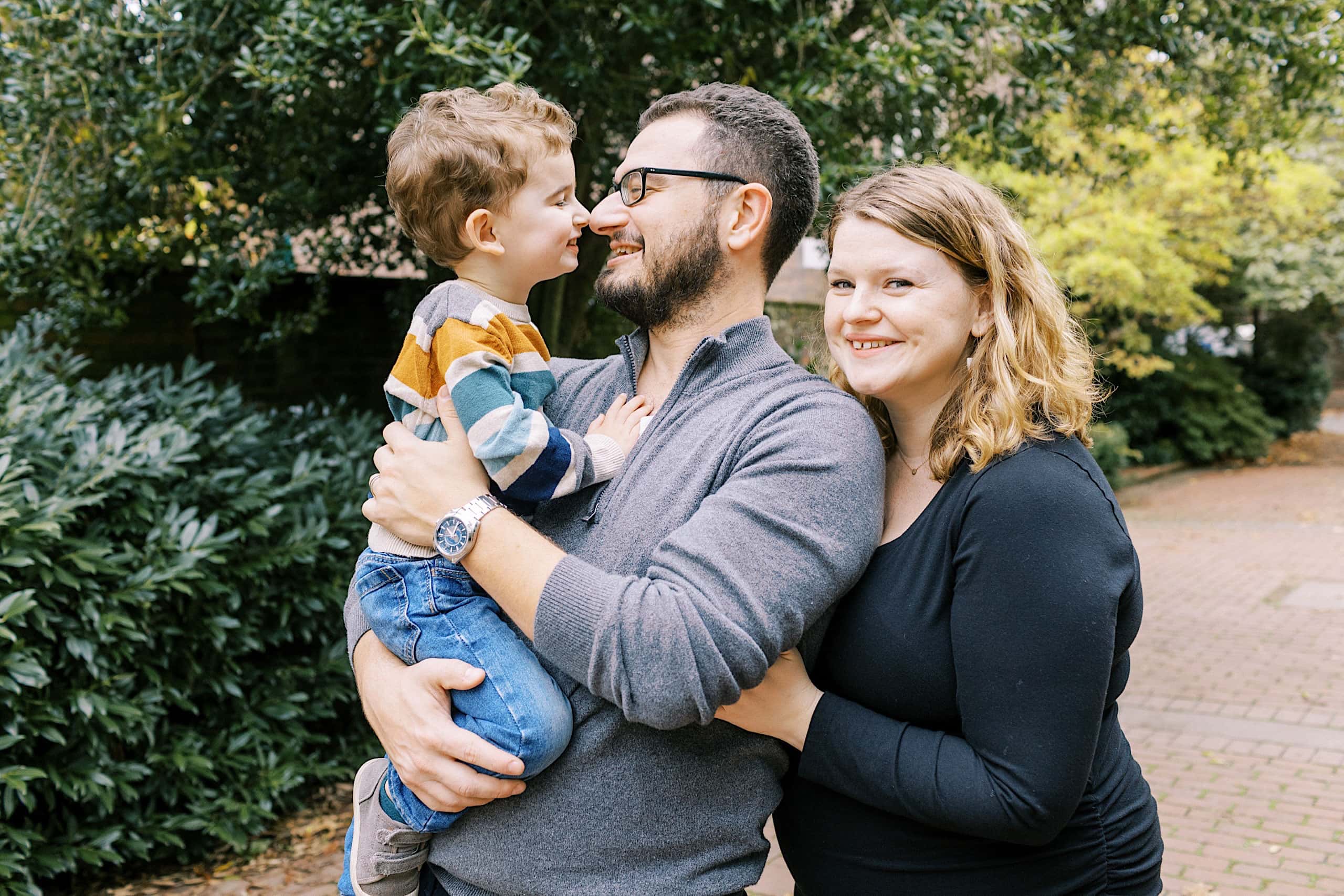 An adult man holds a small boy while a woman stands close beside them. All three are smiling outdoors among green trees and bushes, capturing a joyful moment in these Society Hill Family Photos.