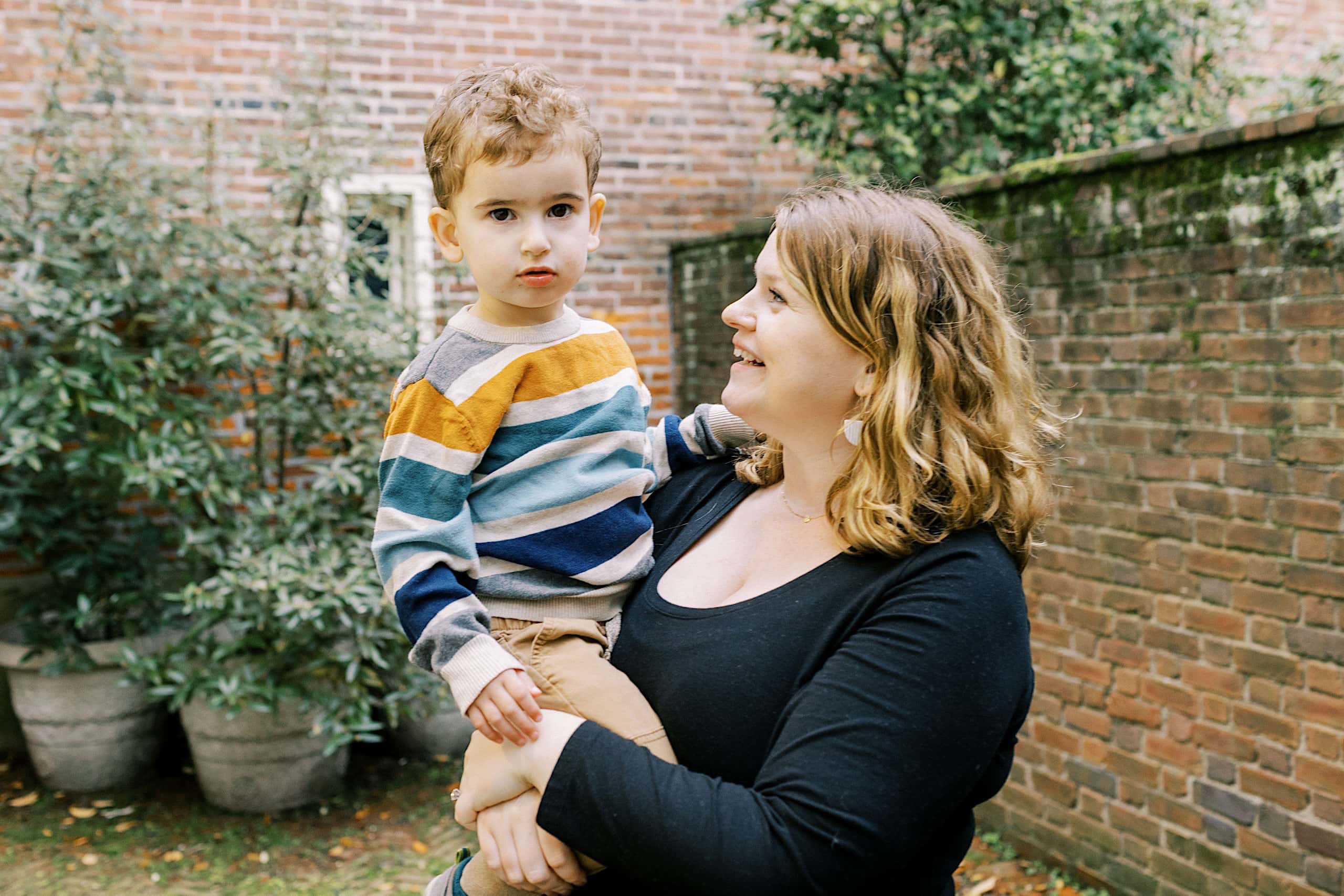 A woman holds a young boy in her arms outdoors near a brick wall and green plants, capturing a warm moment reminiscent of Society Hill Family Photos. The boy looks at the camera while the woman smiles at him.