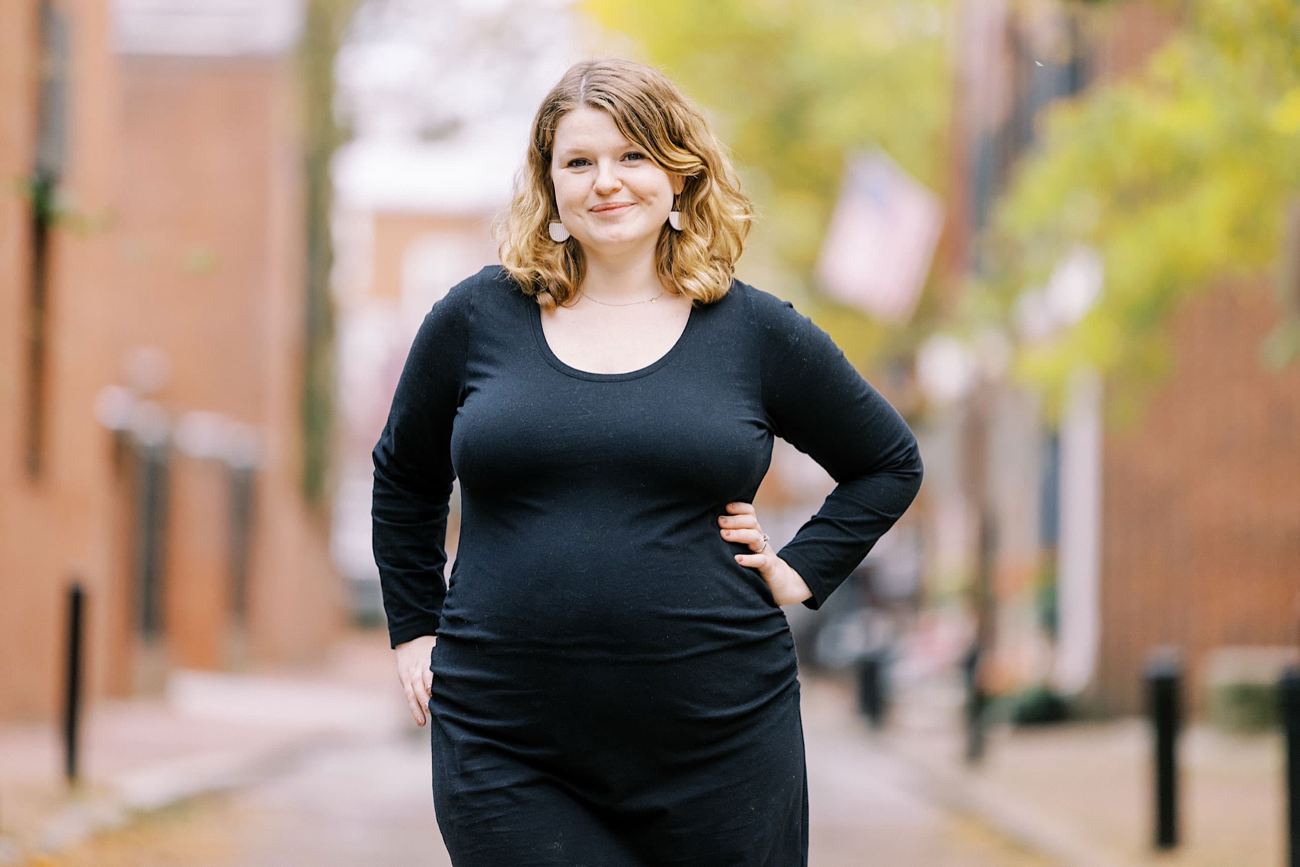 Woman with light brown hair in a black dress stands outdoors on a city street, evoking the timeless charm of Society Hill Family Photos, with trees and classic buildings in the background.