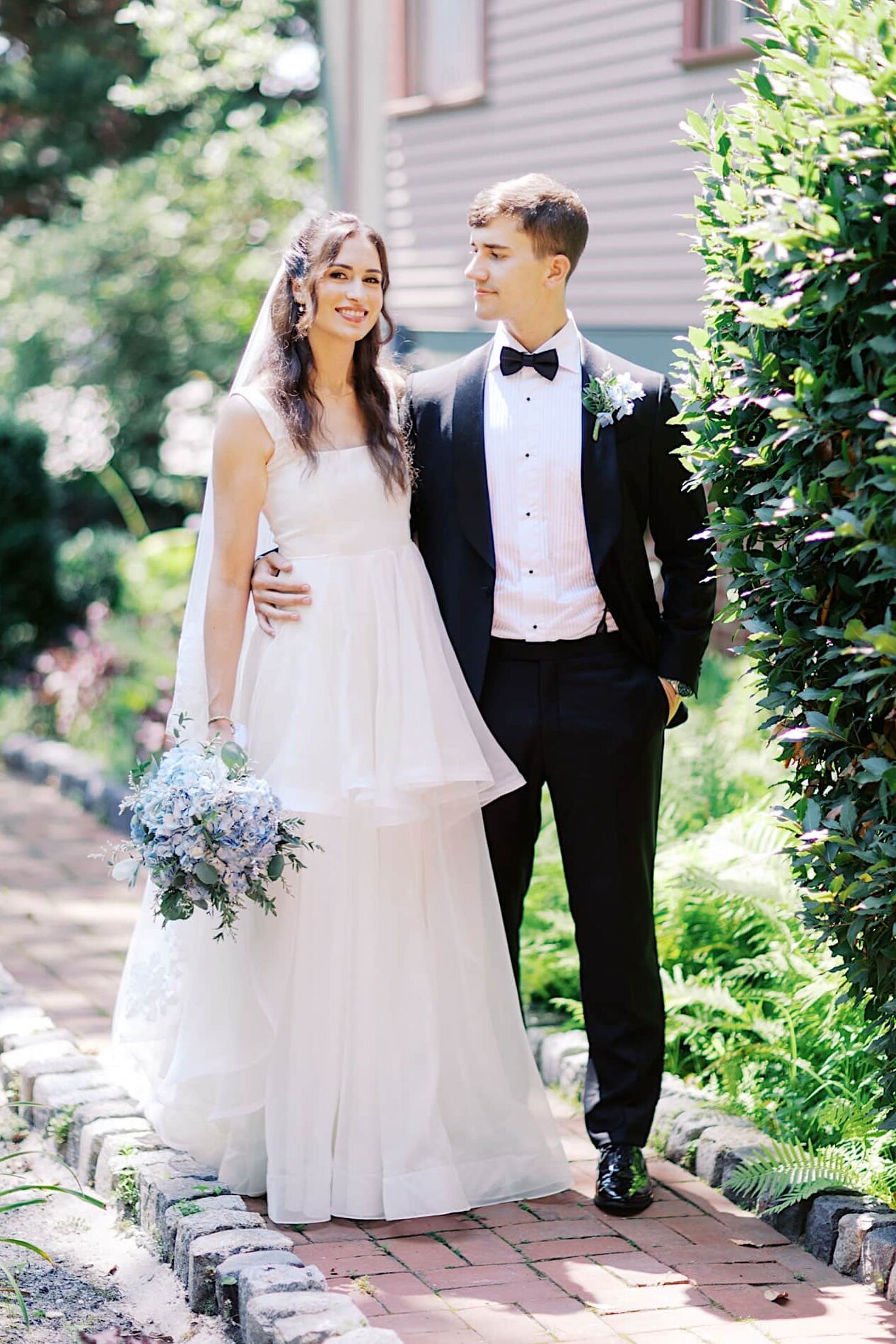 Bride in white dress holding blue hydrangea bouquet; bride and groom standing outdoors in formal attire on a brick path at their elegant Southern Mansion Wedding.