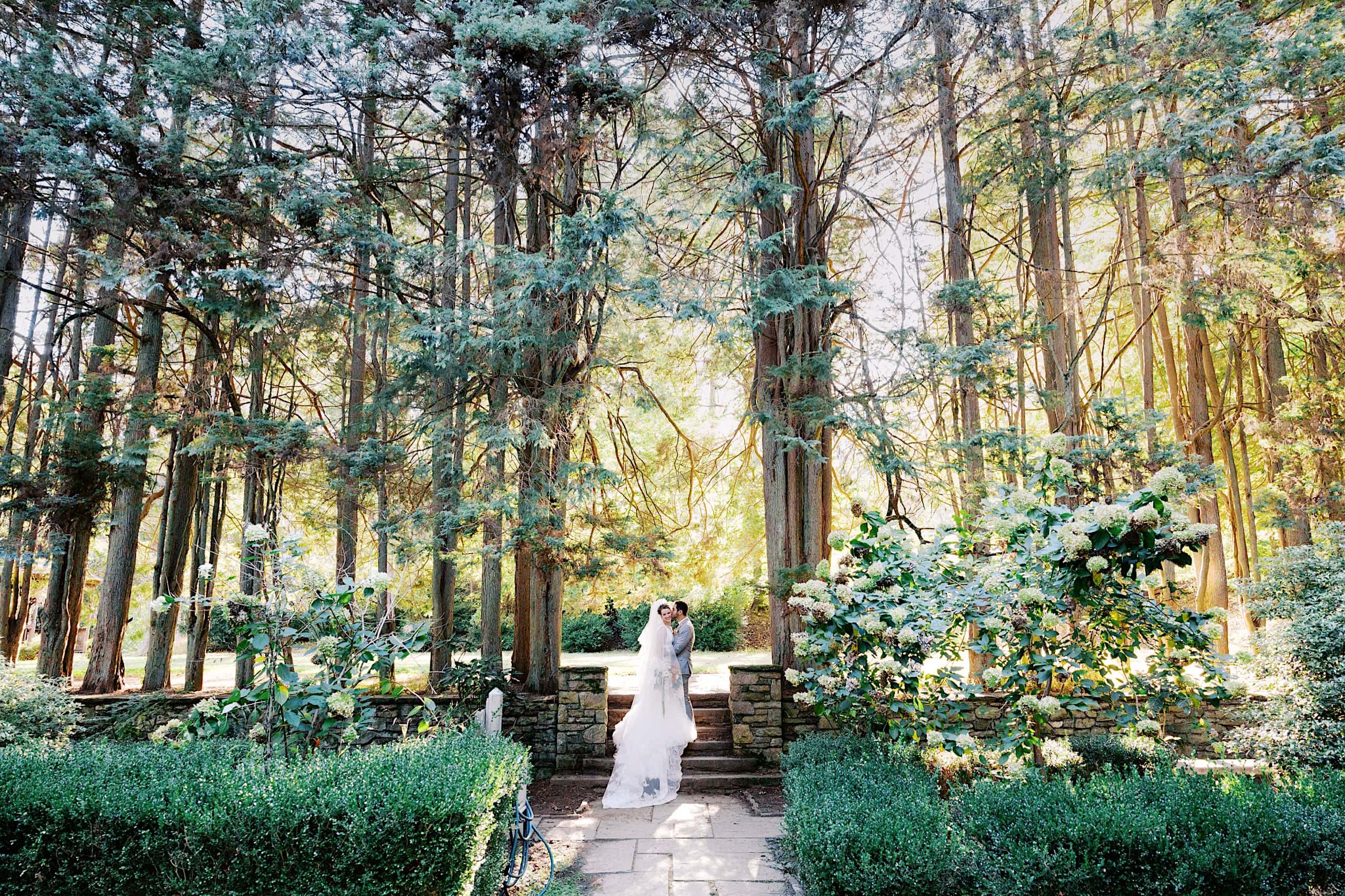 A bride and groom stand together on a stone path in a sunlit forest garden at Parque Ridley Creek, surrounded by tall trees and lush greenery—perfect for an enchanting garden ceremony.