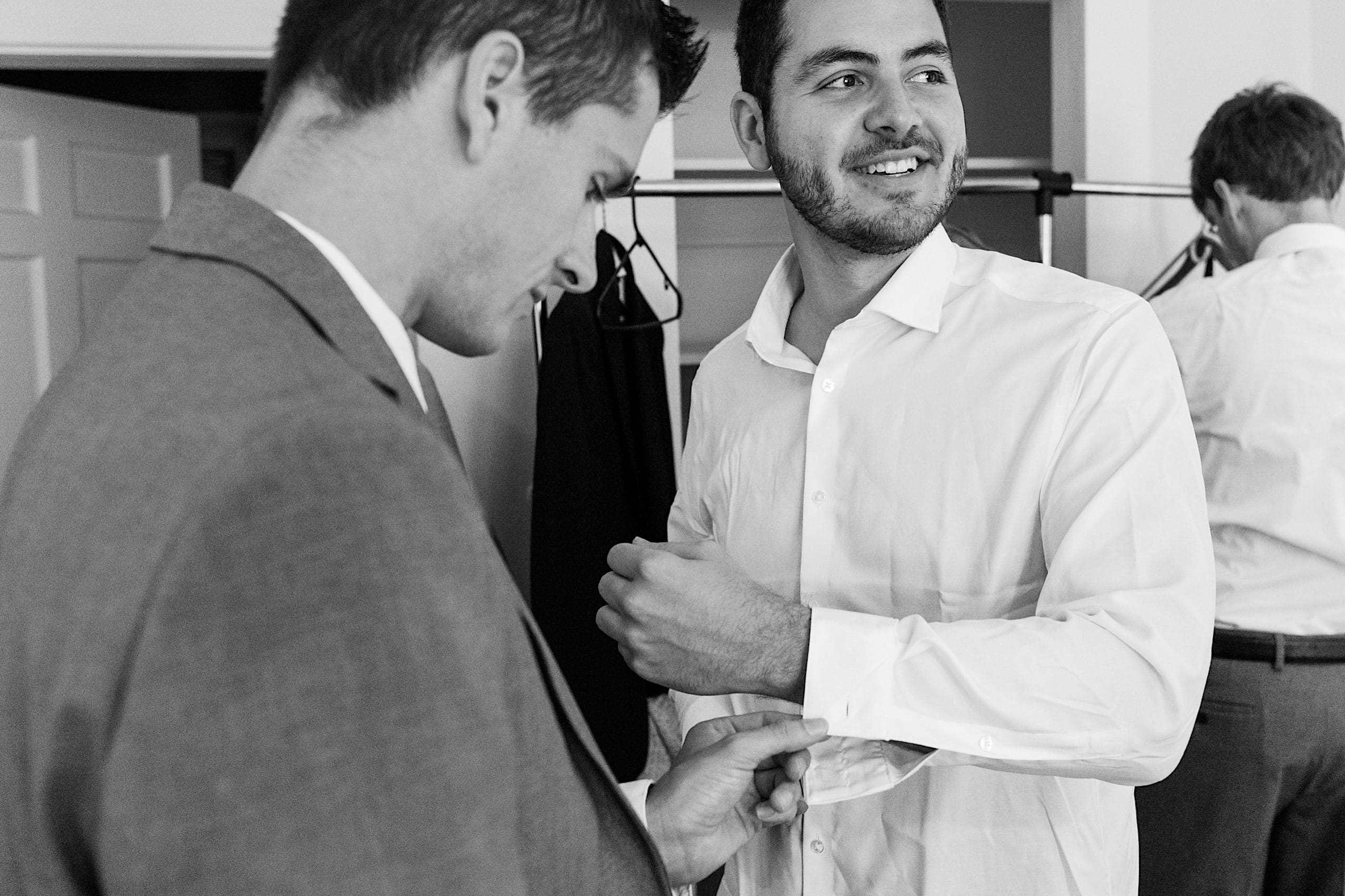 Two men prepare for a formal event—as one adjusts his shirt cuffs, the other assists, both exuding anticipation for the day ahead. In the background, a third man is also getting dressed, all set amid the elegance of a Parque Ridley Creek garden ceremony.