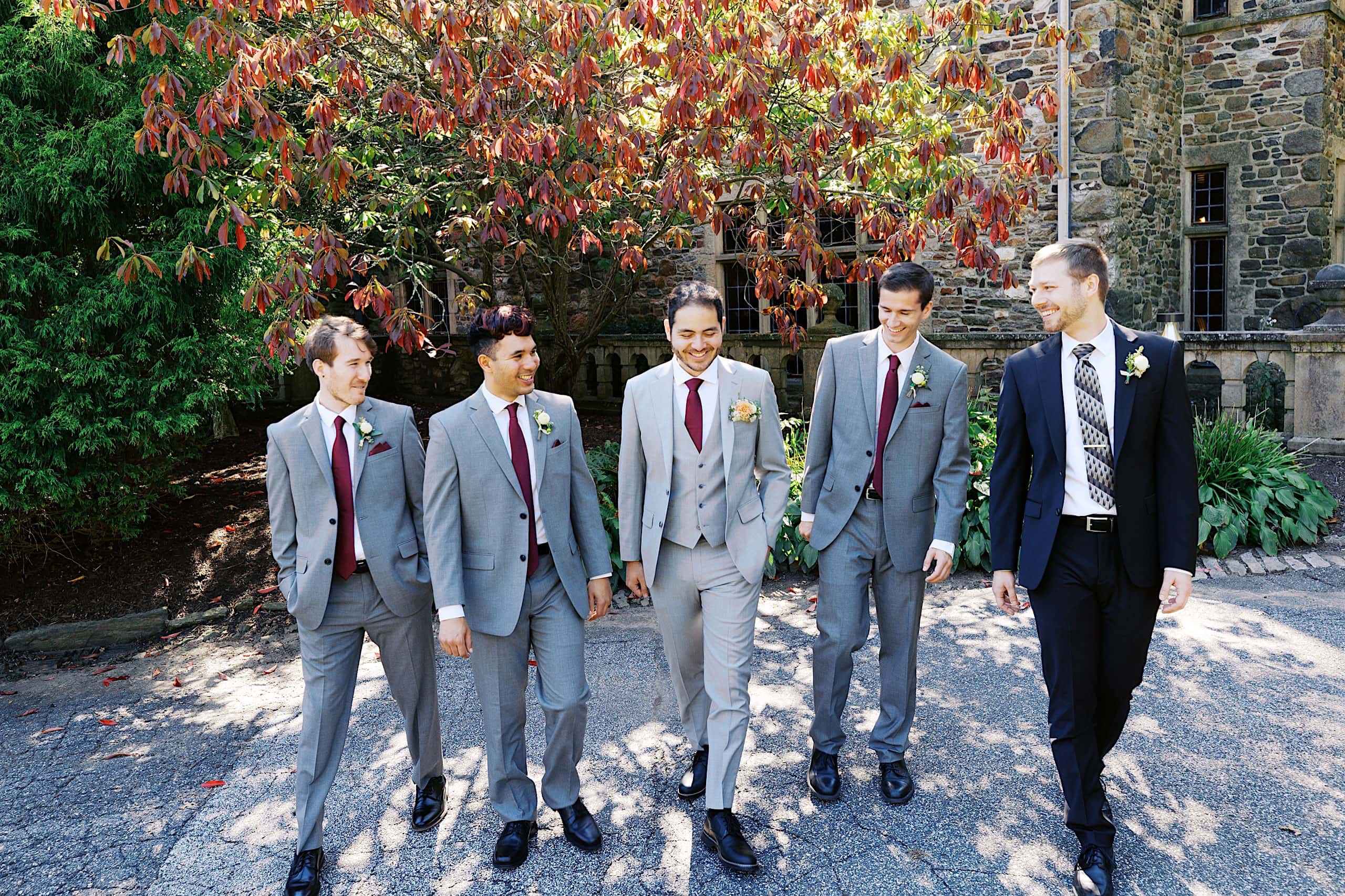 Five men in suits, four in grey and one in black, walk together outside on a sunny day near a stone building and leafy trees, creating an elegant scene reminiscent of a Parque Ridley Creek garden ceremony.