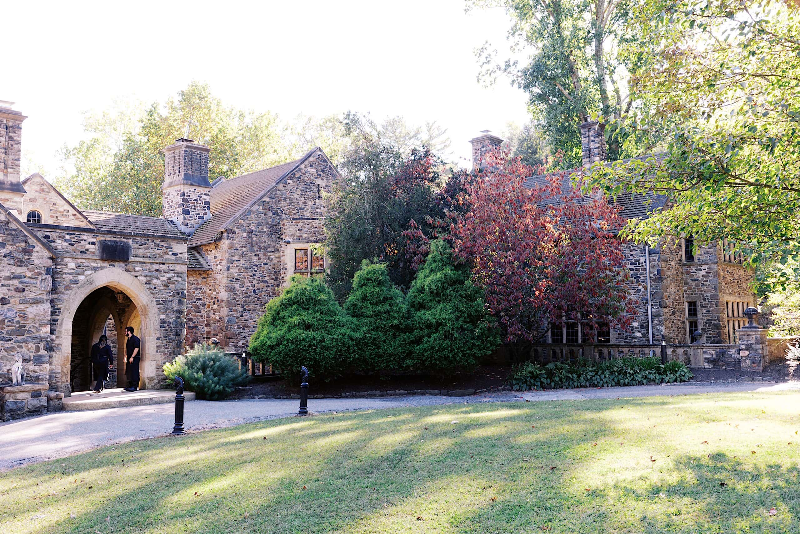 A stone building with arched entryway, multiple chimneys, and leafy trees surrounding the structure on a sunny day—perfect for a Parque Ridley Creek garden ceremony.
