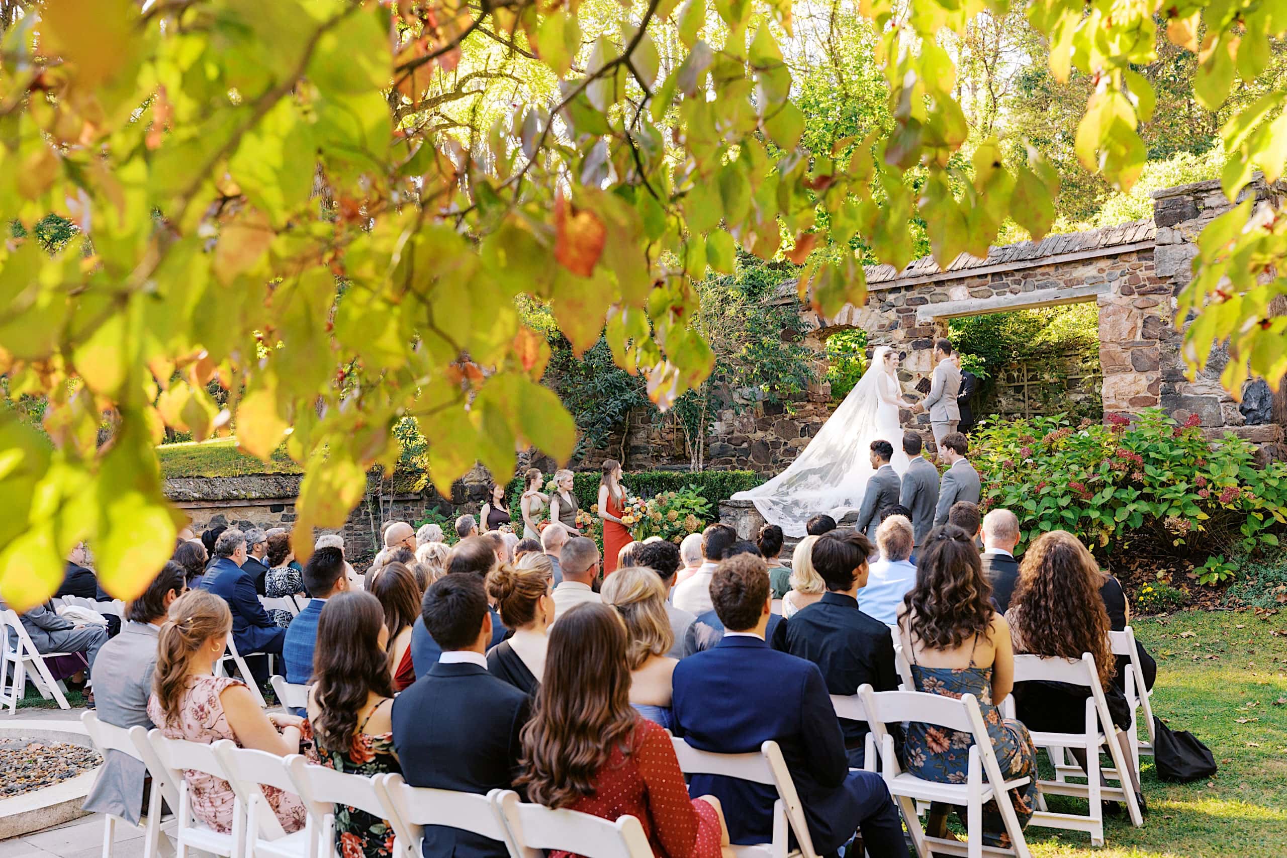 Outdoor wedding ceremony with guests seated on white chairs, bride and groom standing before an officiant near a stone arch—this Parque Ridley Creek garden ceremony is surrounded by lush greenery and sunlight gently filtering through the trees.