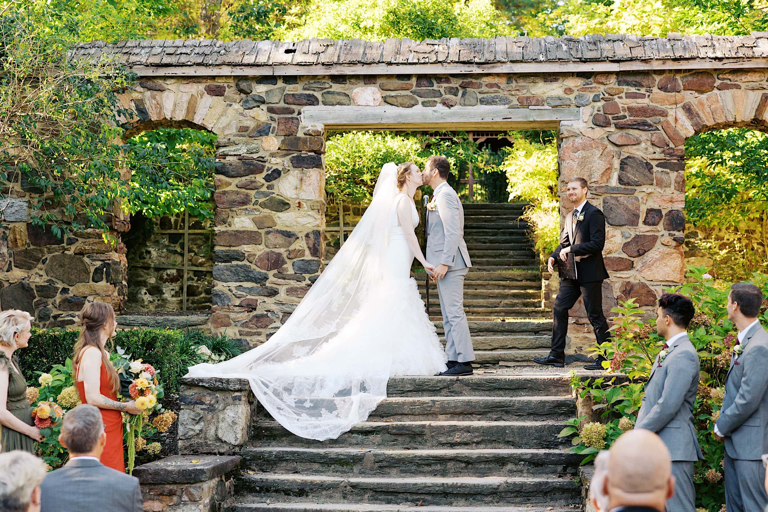 A bride and groom kiss at the top of stone steps during an outdoor wedding ceremony in the Parque Ridley Creek garden, surrounded by their wedding party and officiant, with greenery and sunlight in the background.