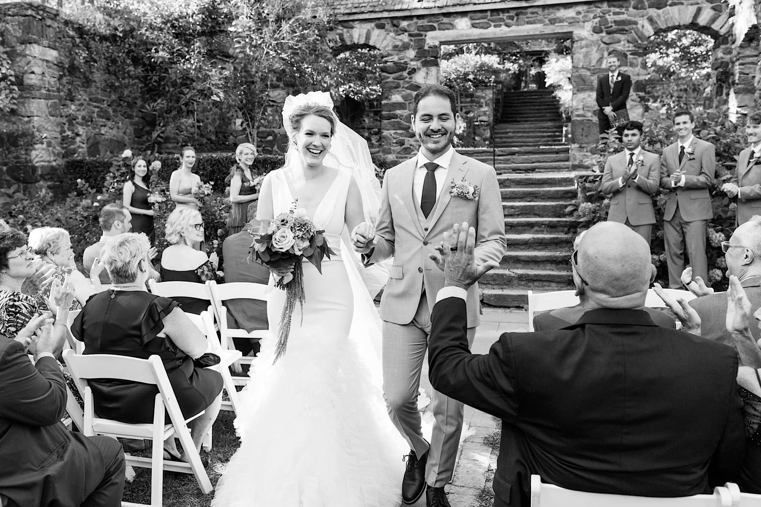A bride and groom walk down the aisle outdoors during their Parque Ridley Creek garden ceremony, smiling as guests seated in white chairs clap and celebrate their wedding.