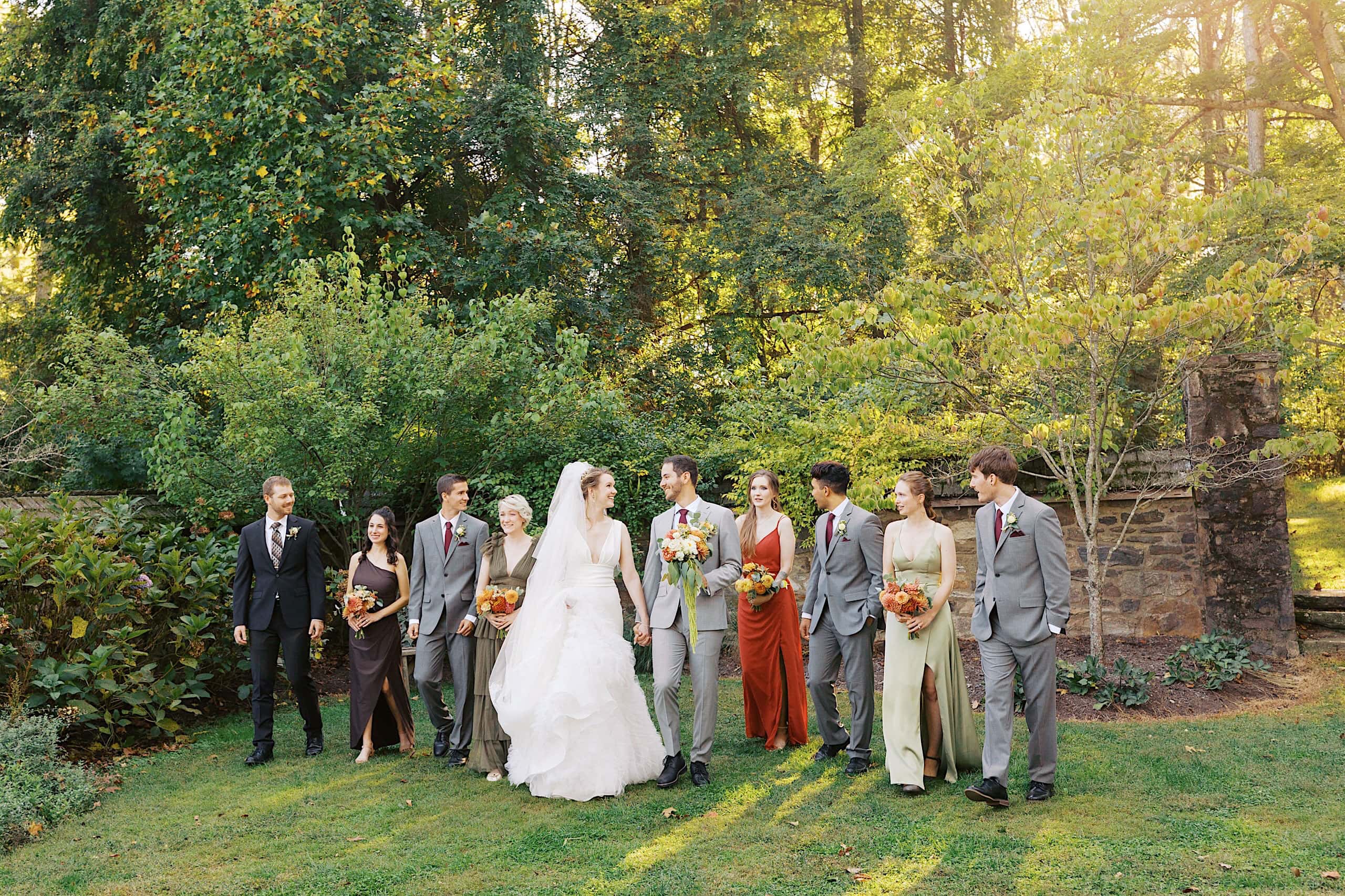 A bride and groom walk outdoors with their wedding party, all dressed formally, surrounded by trees and greenery on a sunny day, following a beautiful Parque Ridley Creek garden ceremony.