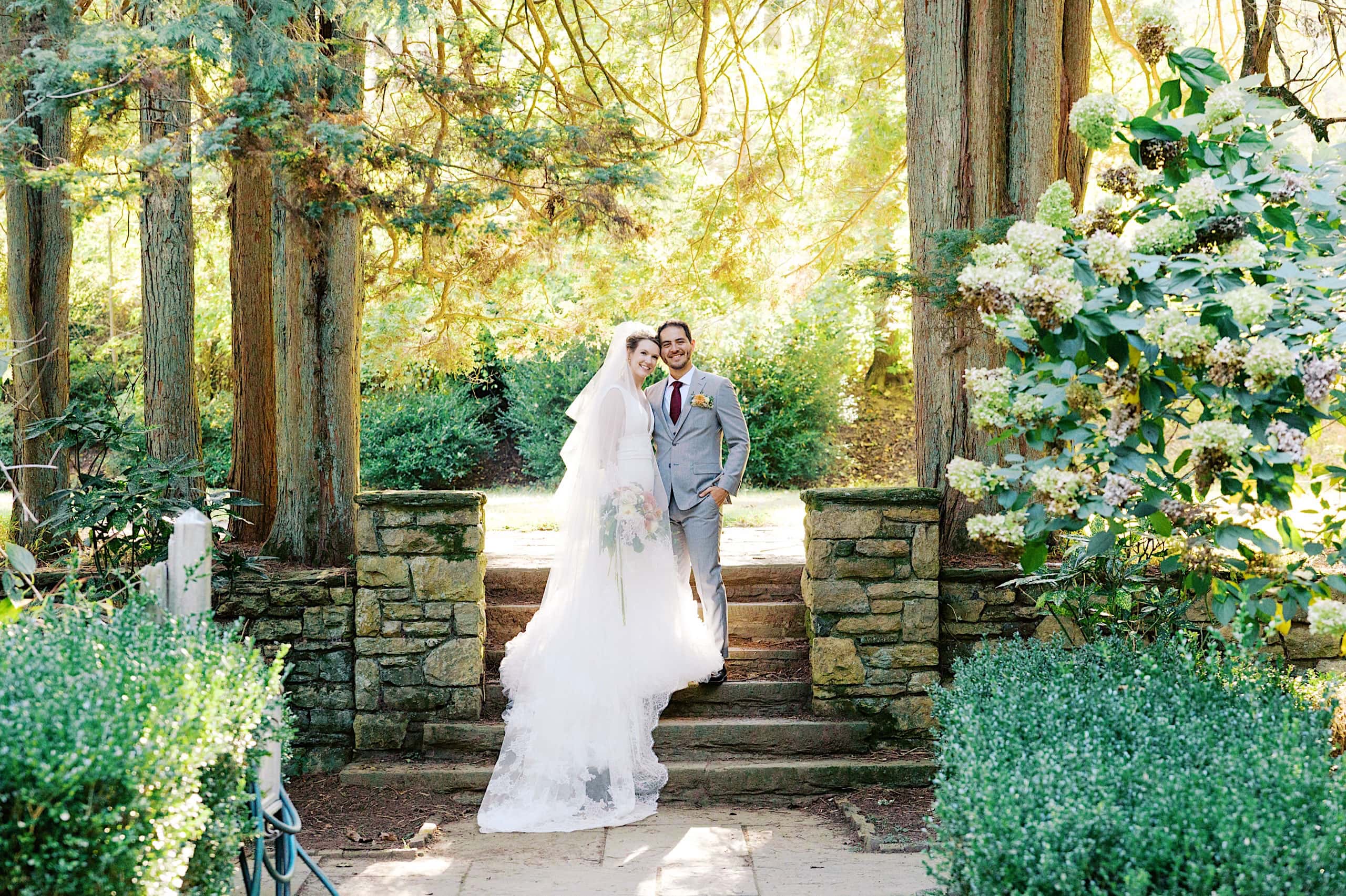 A bride in a white dress and veil stands beside a groom in a light gray suit, posing outdoors among trees and stone walls on a sunny day, capturing the romance of their Parque Ridley Creek garden ceremony.