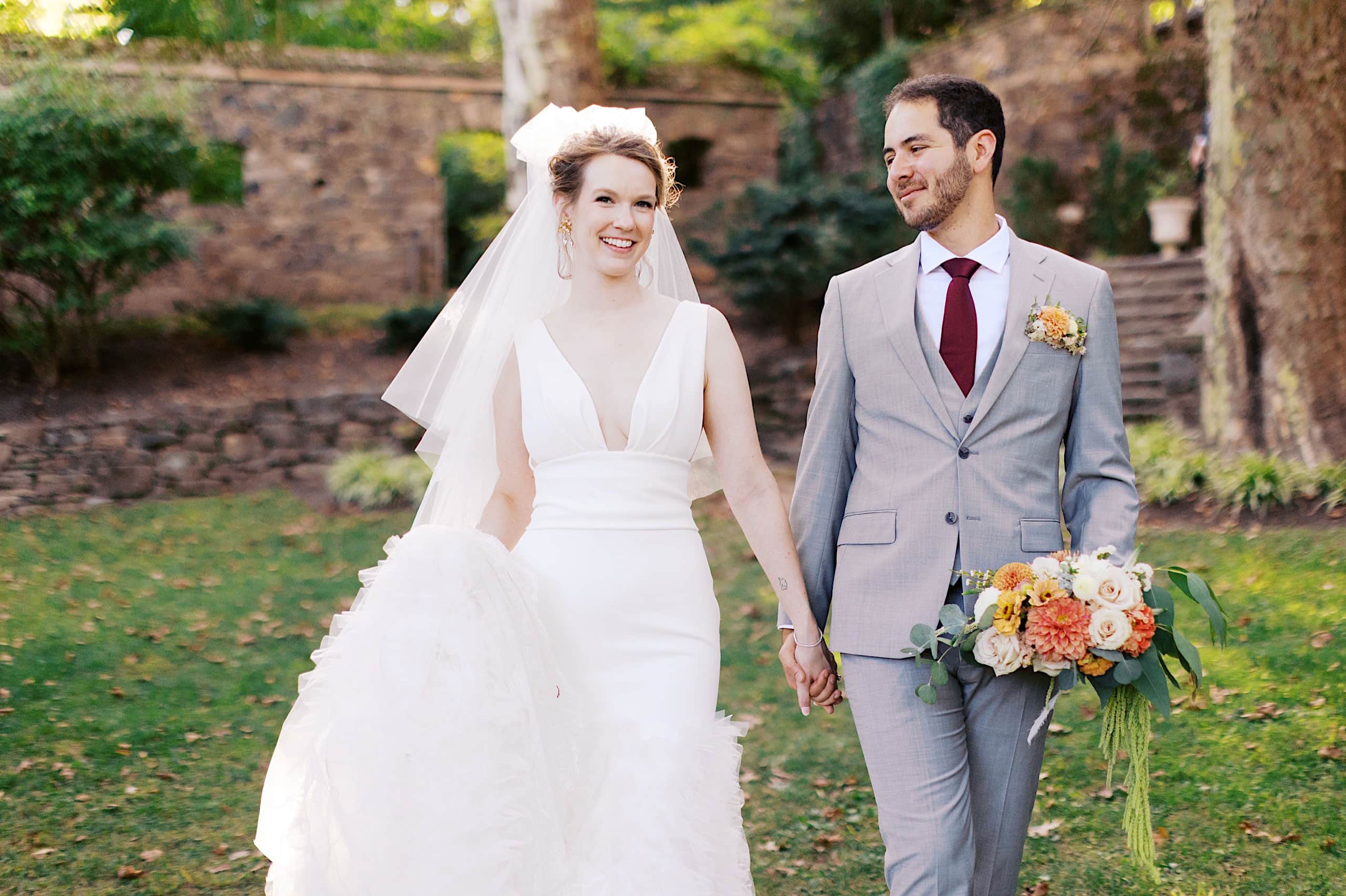 A bride in a white dress and veil walks hand-in-hand with a groom in a light gray suit holding a bouquet, outdoors during their Parque Ridley Creek garden ceremony.