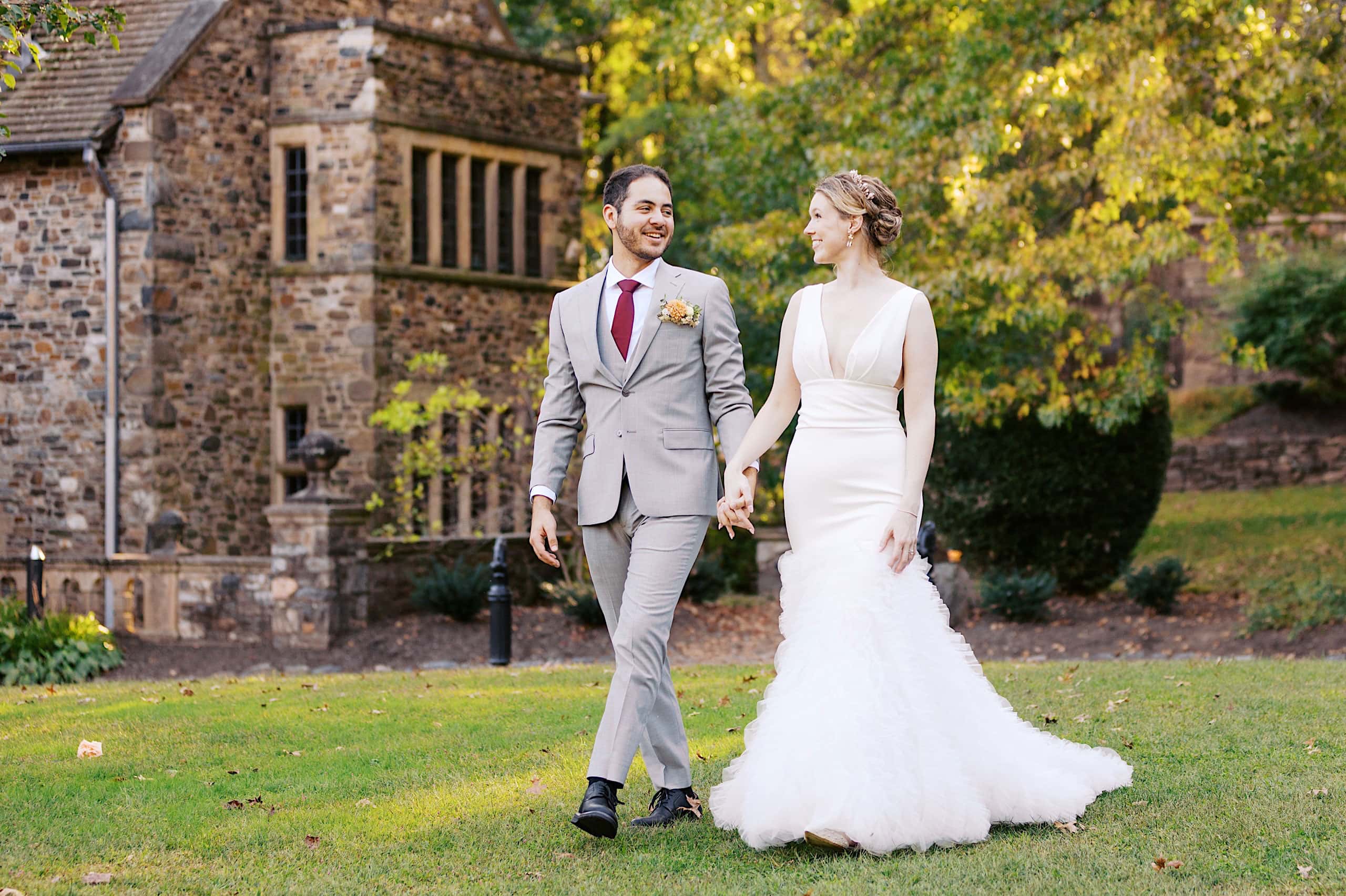 A couple dressed in wedding attire walks hand in hand on grass near a stone building with large windows and trees in the background, capturing the enchanting atmosphere of a Parque Ridley Creek garden ceremony.