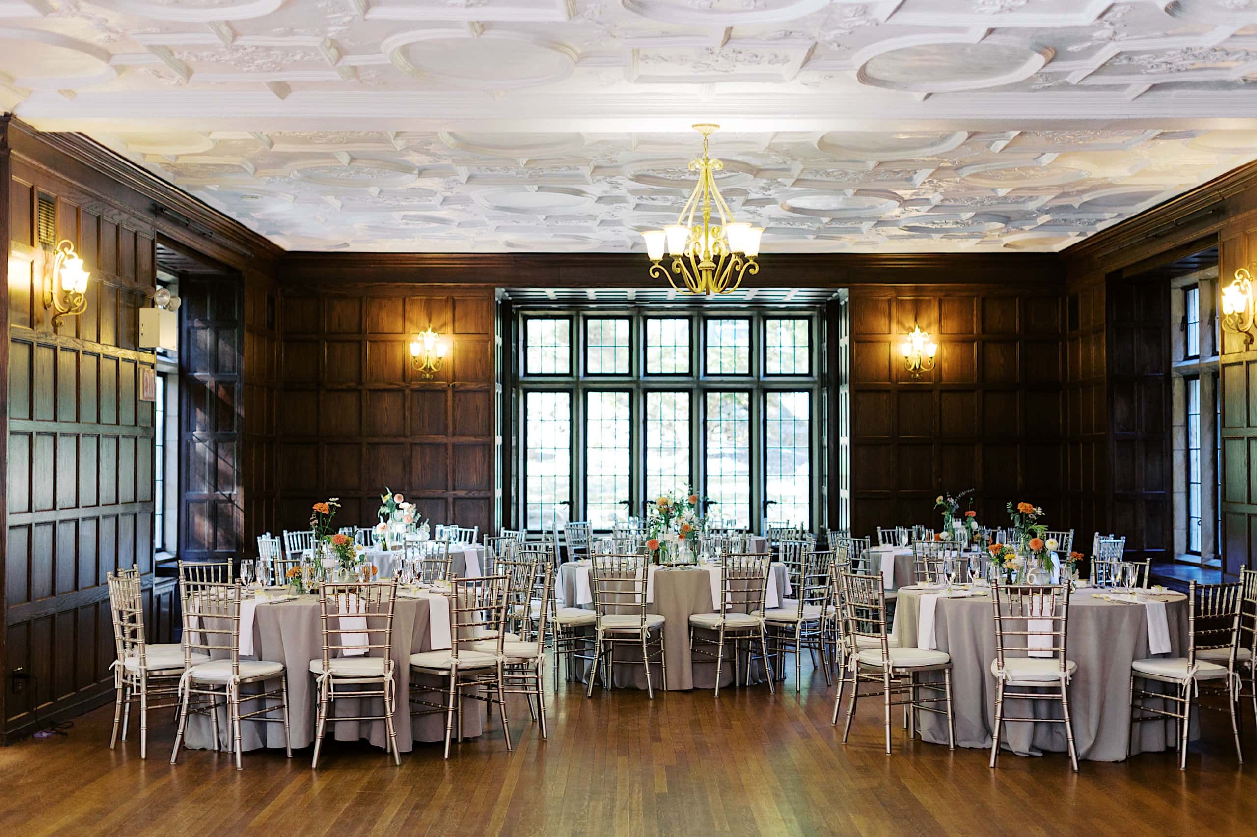 Elegant banquet hall with wood-paneled walls, round tables set with flowers, and silver chairs arranged for an event under ornate ceiling lights—an ideal indoor complement to a Parque Ridley Creek garden ceremony.