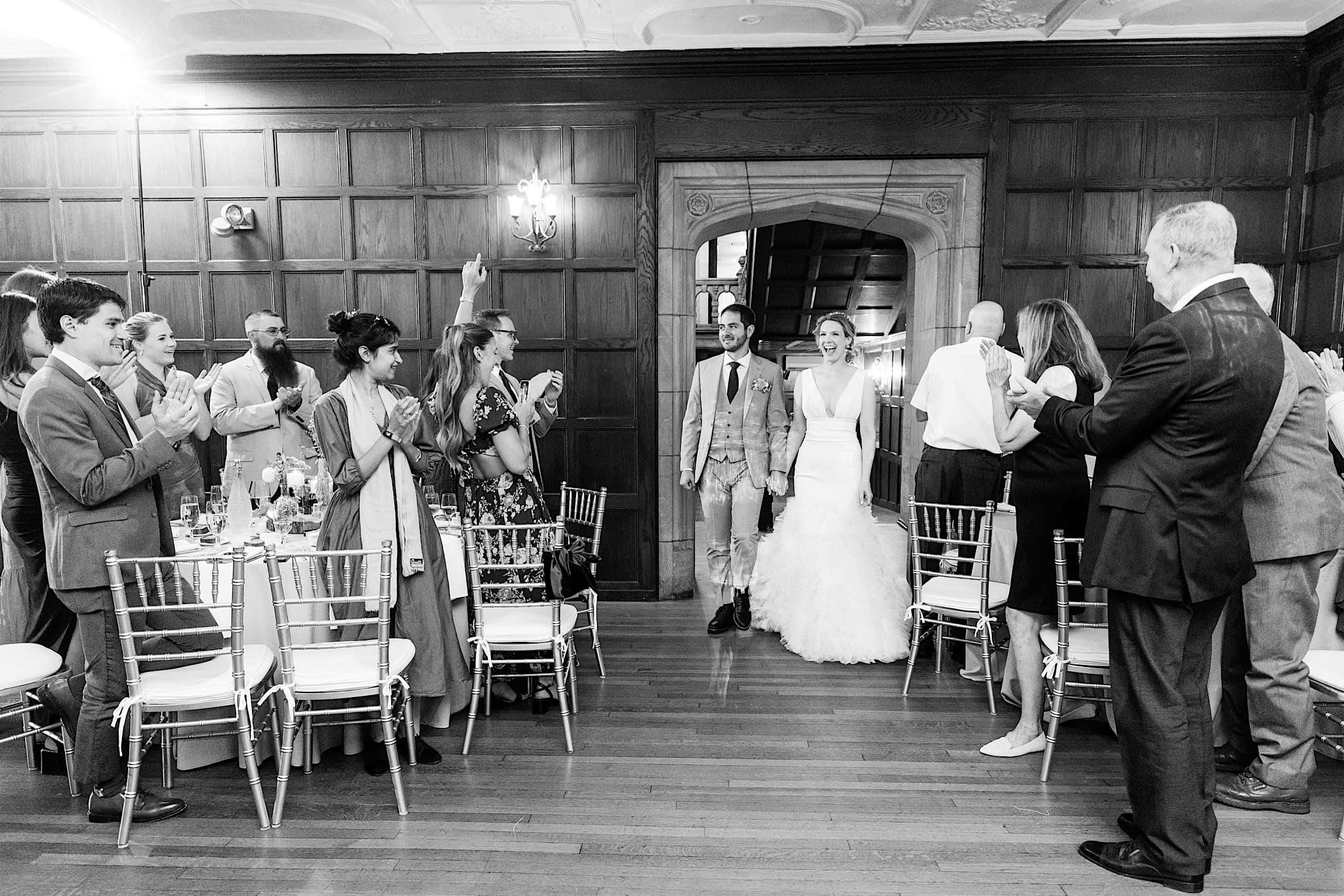 A bride and groom enter a wood-paneled room as guests stand and applaud, some holding up phones to take photos, still glowing from their Parque Ridley Creek garden ceremony.
