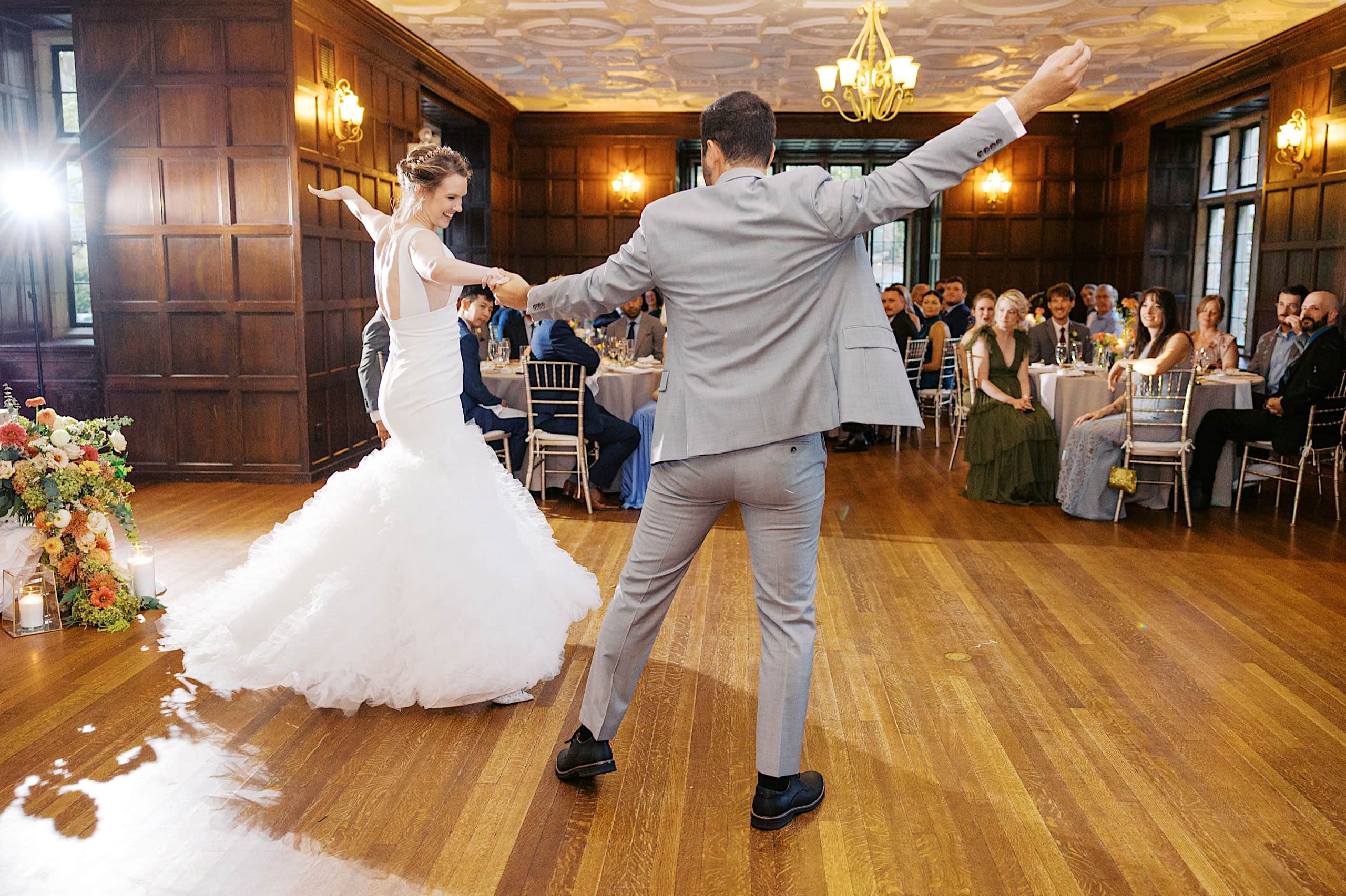 A bride and groom dance together in a wood-paneled room as wedding guests seated at round tables watch, their celebration following a beautiful Parque Ridley Creek garden ceremony.