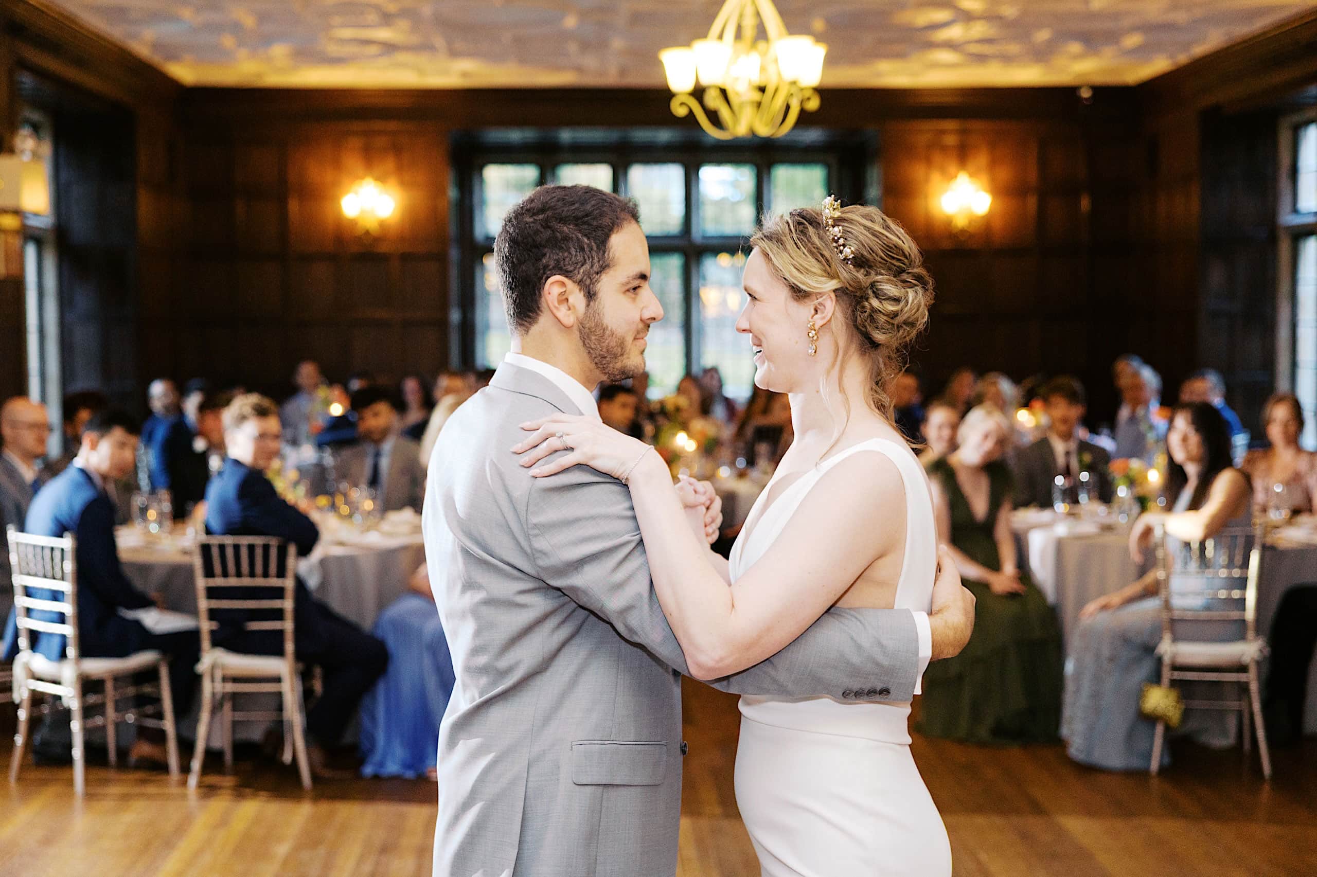 A bride and groom dance together in a wood-paneled room as seated guests watch from round tables in the background, celebrating after their romantic Parque Ridley Creek garden ceremony.