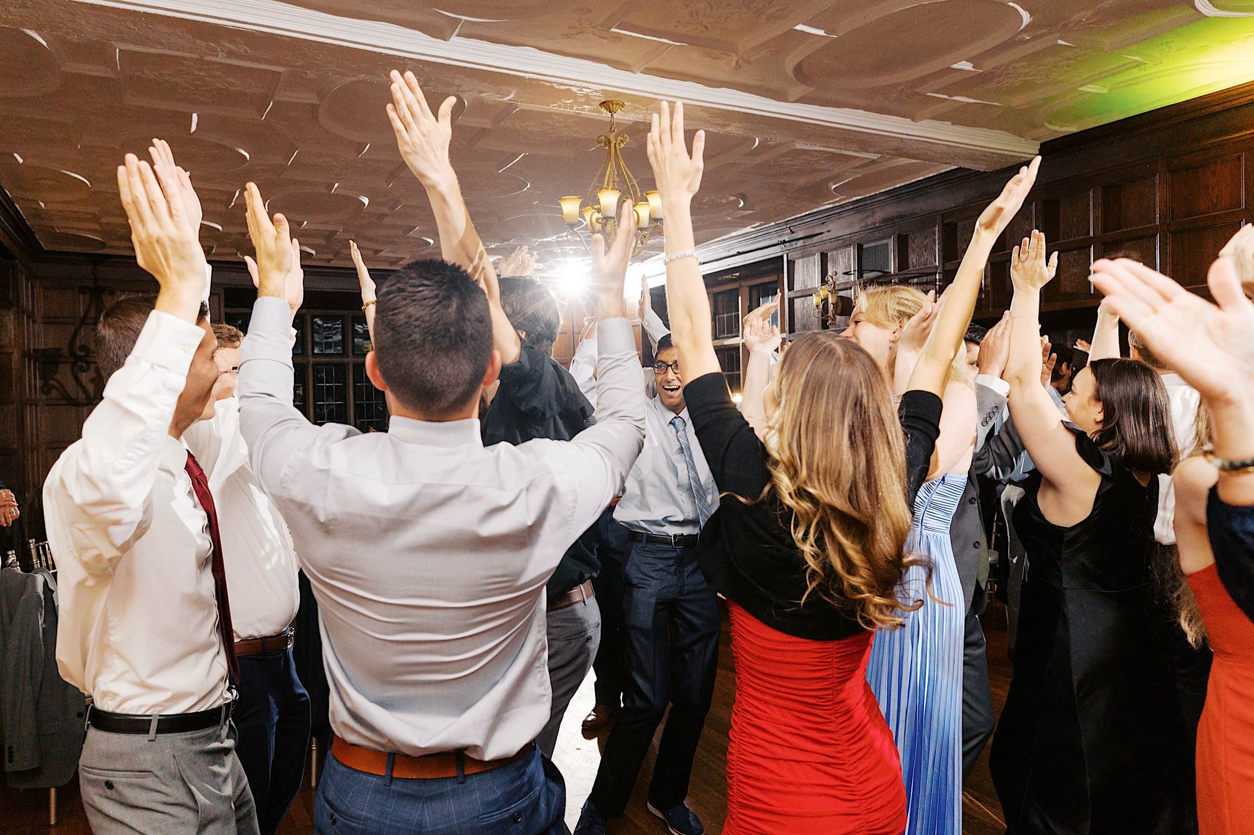A group of people dressed formally stand in a circle with their arms raised, appearing to celebrate or dance together as if recreating the joy of a Parque Ridley Creek garden ceremony within the warmth of a wood-paneled room.