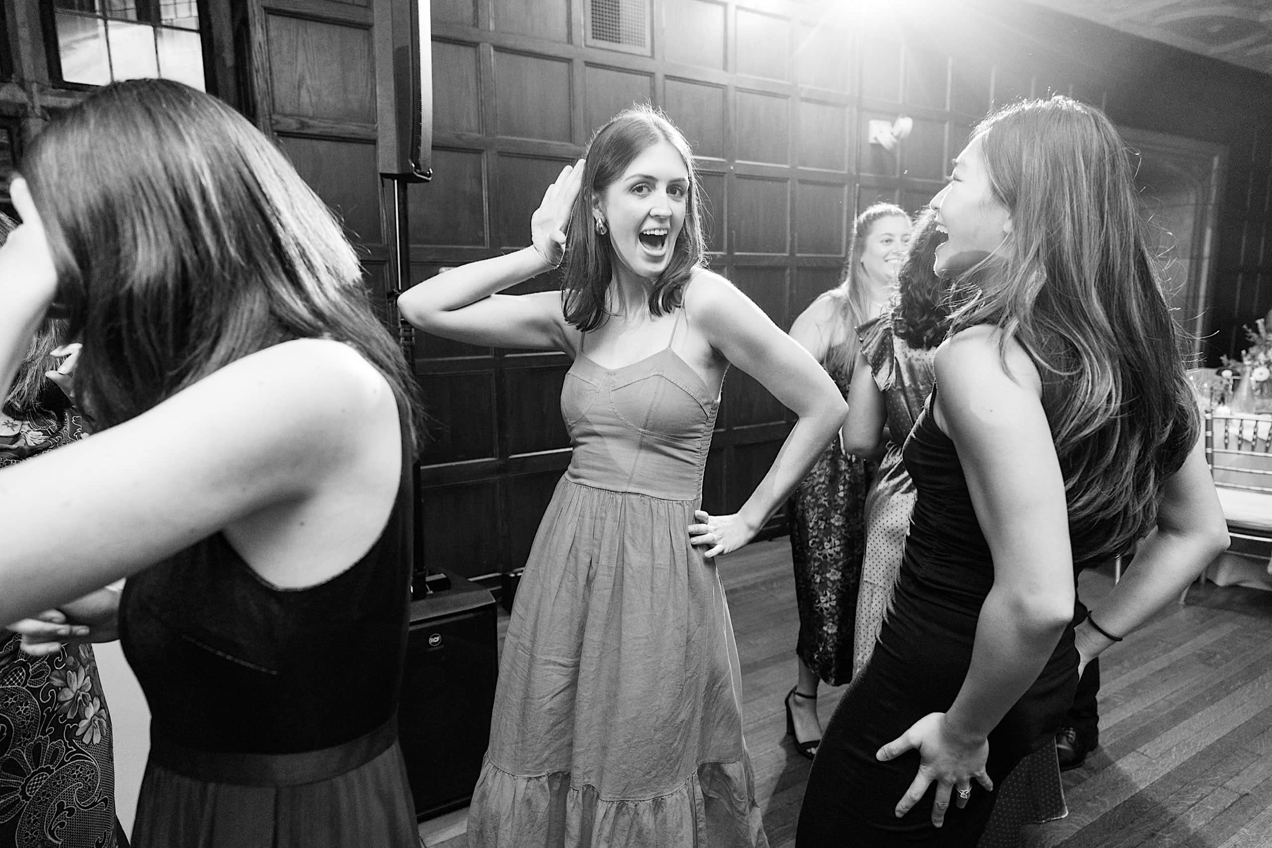 A group of women in dresses dance and interact at an indoor event reminiscent of a Parque Ridley Creek garden ceremony, with one woman in the center holding her hand to her ear, smiling.