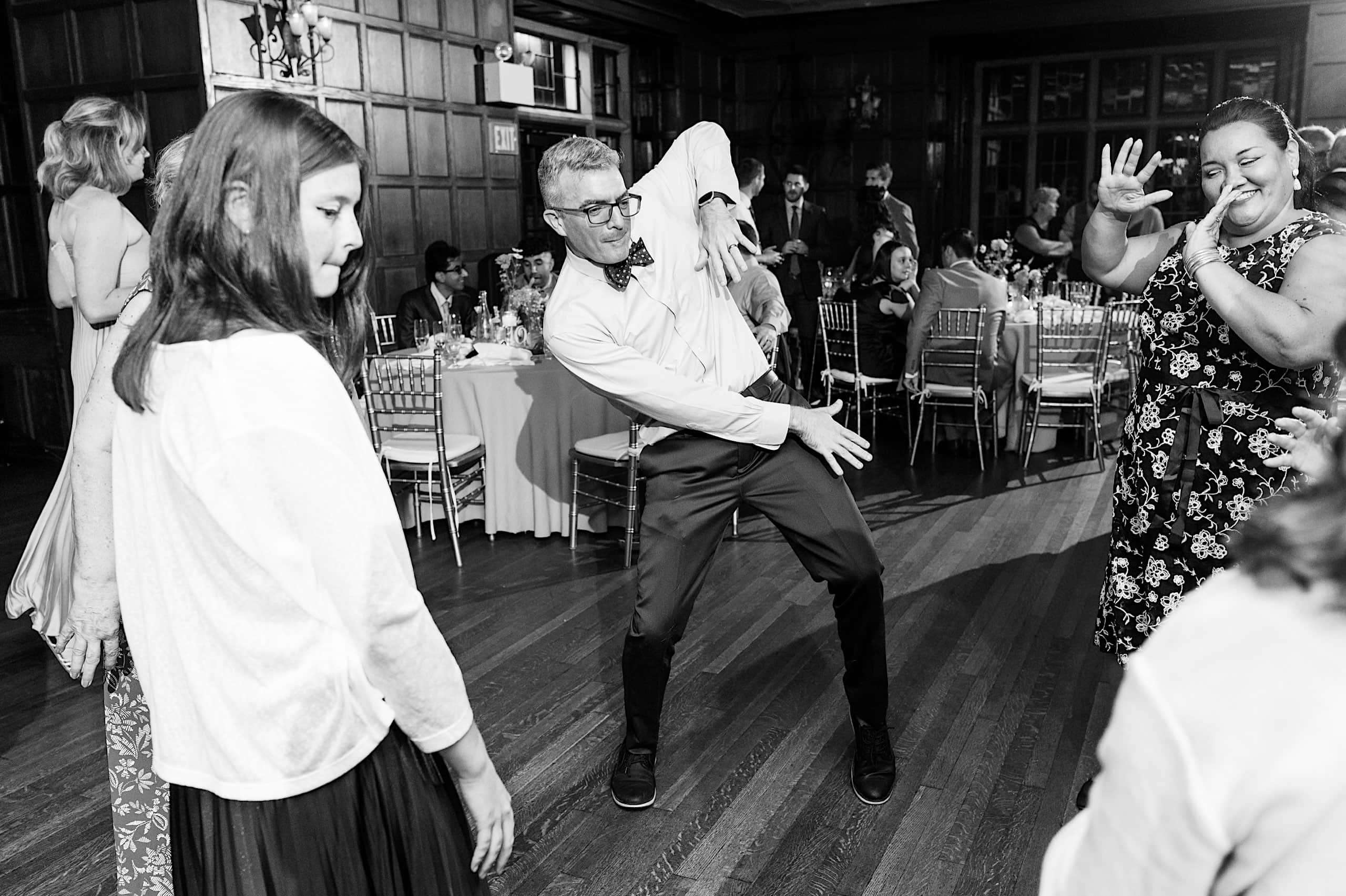 A group of people dance in a formal room with wooden walls and banquet tables, recalling the elegance of a Parque Ridley Creek garden ceremony; one man poses energetically while others watch and smile.