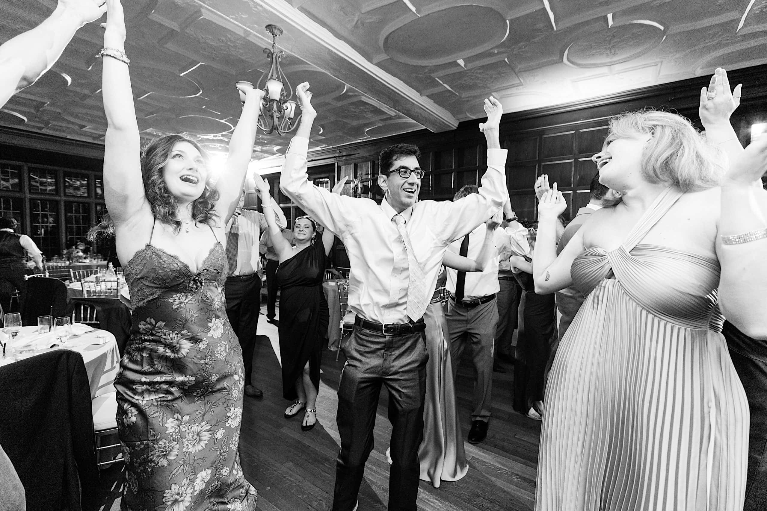 A group of people in formal attire dance with their arms raised at an indoor event with wooden paneling and a decorative ceiling, evoking the joyful spirit of a Parque Ridley Creek garden ceremony.