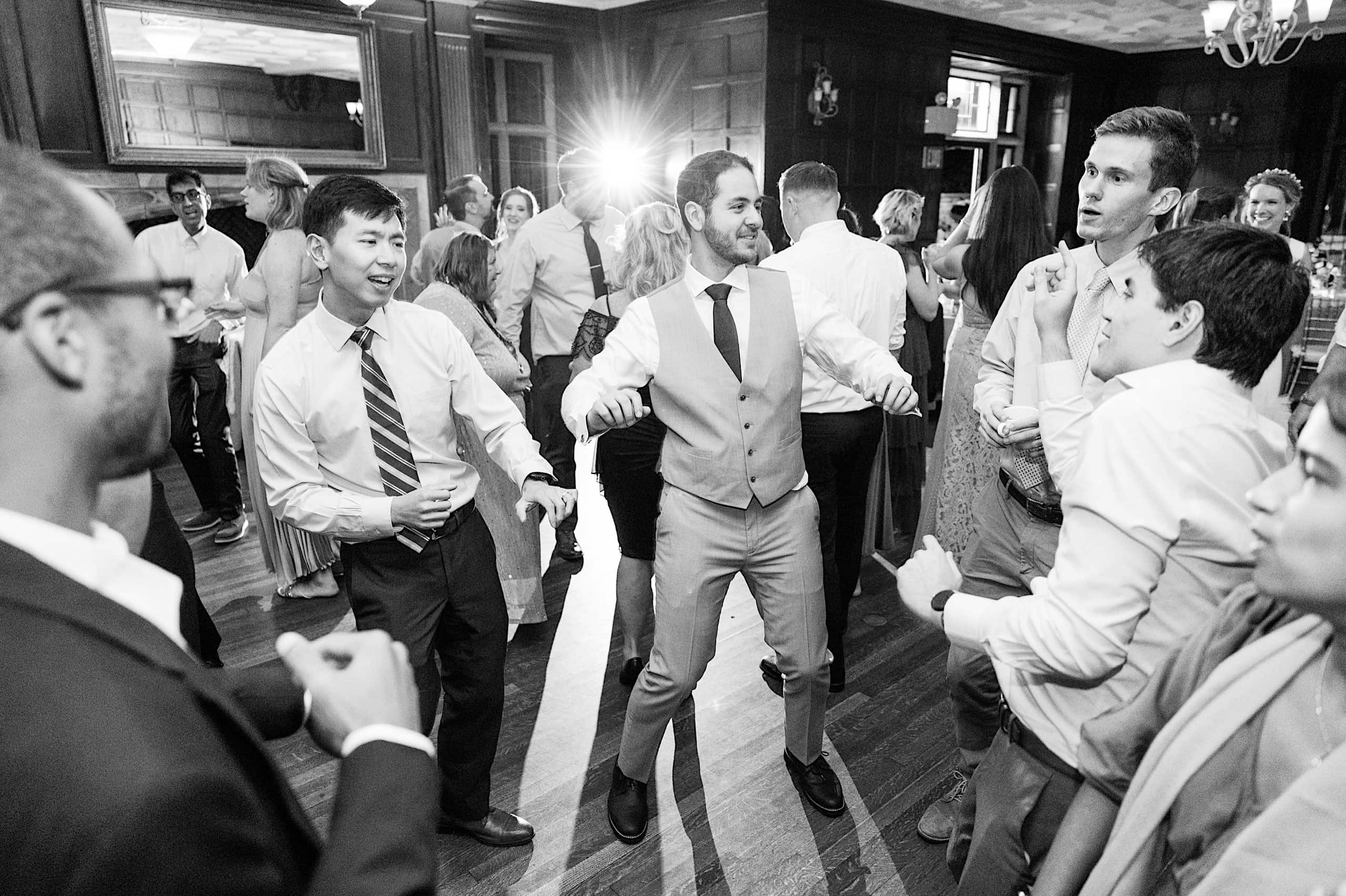 A group of men in dress shirts and suits dance and talk at an indoor event, reminiscing about the beautiful Parque Ridley Creek garden ceremony earlier in the day, with other guests and a bright light visible in the background.