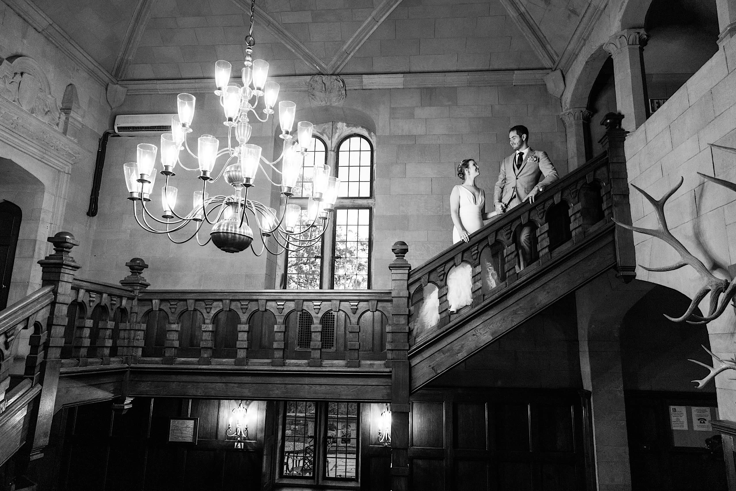 A bride and groom stand on a grand wooden staircase in a historic building with high ceilings, large windows, and a chandelier, reminiscing about their Parque Ridley Creek garden ceremony.