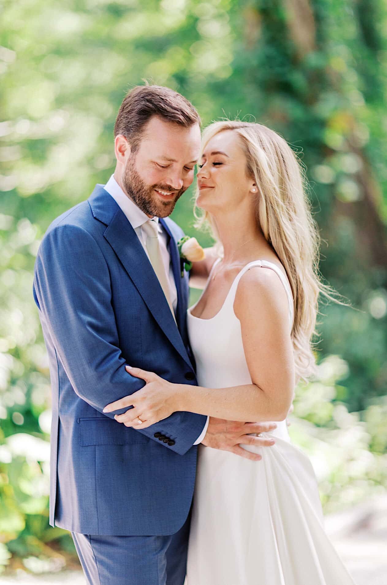 A bride in a white gown and a groom in a blue suit stand together in a wooded area of Beach Plum Farm on their wedding day.