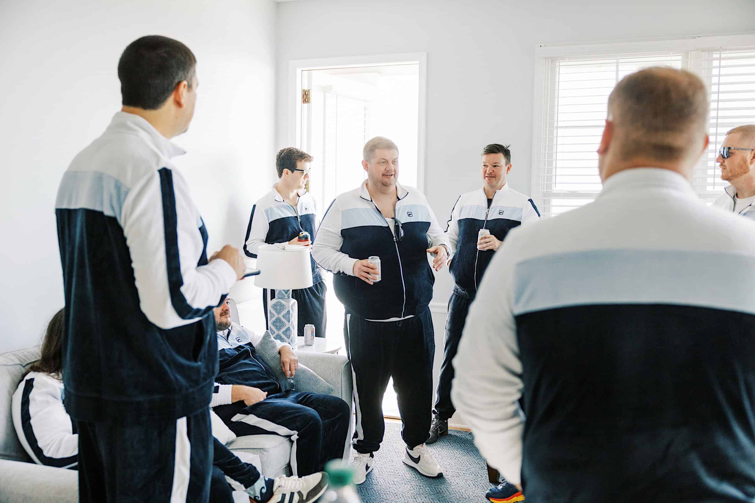 A group of men in matching tracksuits stand and sit in a bright room, talking and holding drinks, preparing for a romantic November wedding at Congress Hall.