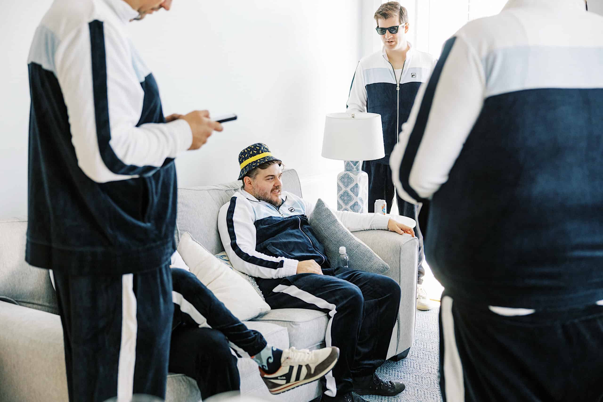 Four men in matching tracksuits are gathered in a bright, modern living room. One, wearing a party hat, sits on the sofa as others stand nearby—perhaps reminiscing about a romantic November wedding at Congress Hall.