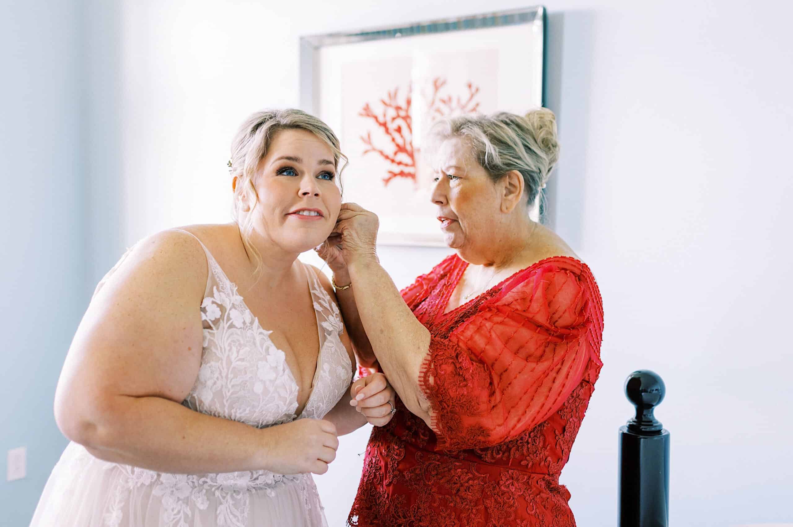 A woman in a white lace dress has her earring adjusted by an older woman in red, standing together in a brightly lit room with pale blue walls before a romantic November wedding at Congress Hall.