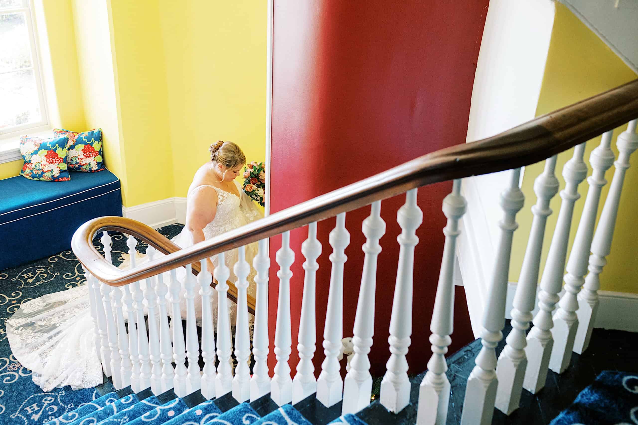 A bride in a white gown holds a bouquet while walking up a blue-carpeted staircase with white railings during a romantic November wedding at Congress Hall in a brightly lit interior.