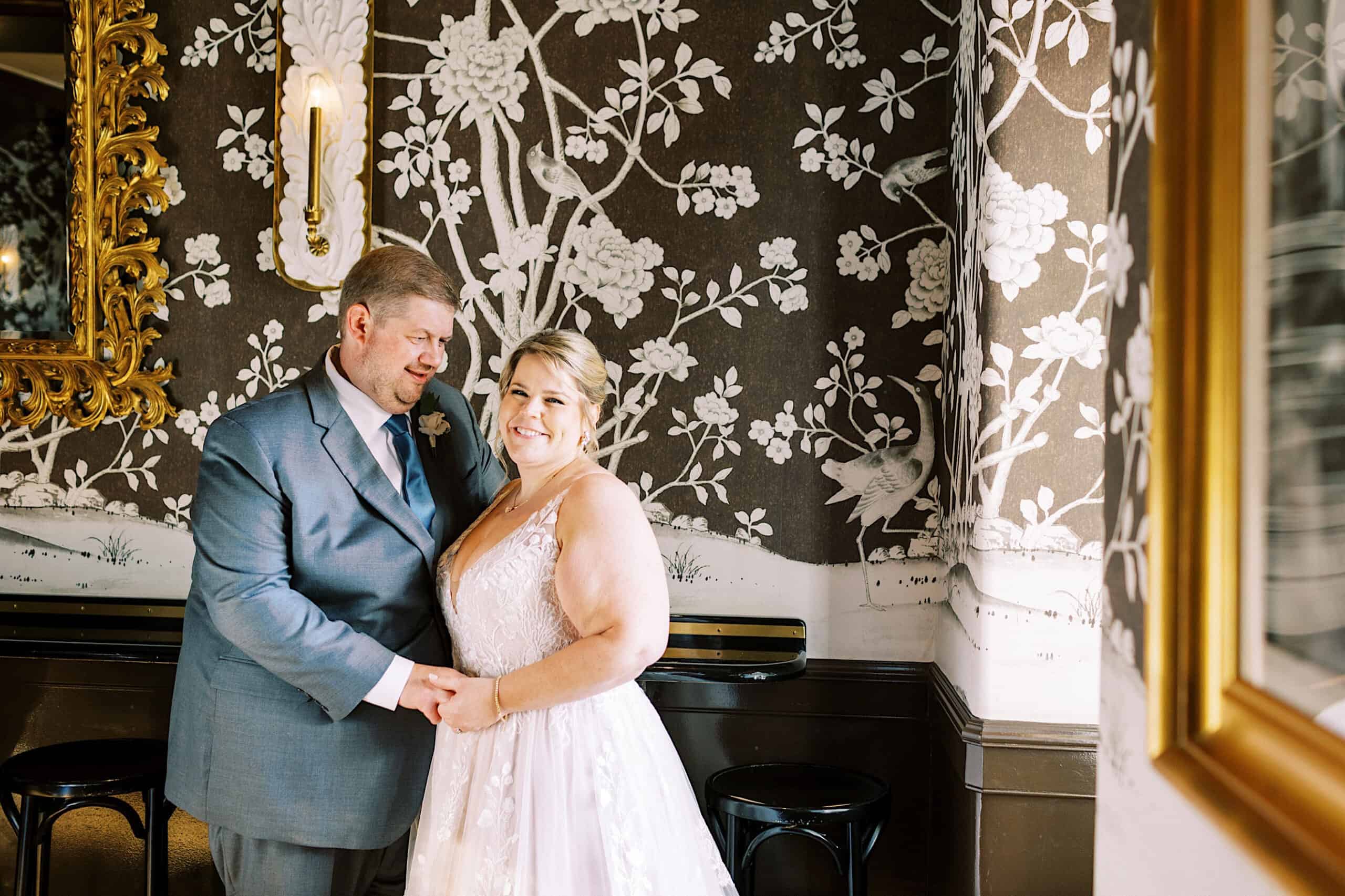 A couple dressed in wedding attire stands together, smiling and holding hands in a room with floral-patterned wallpaper and gold-framed mirrors during their romantic November wedding at Congress Hall.