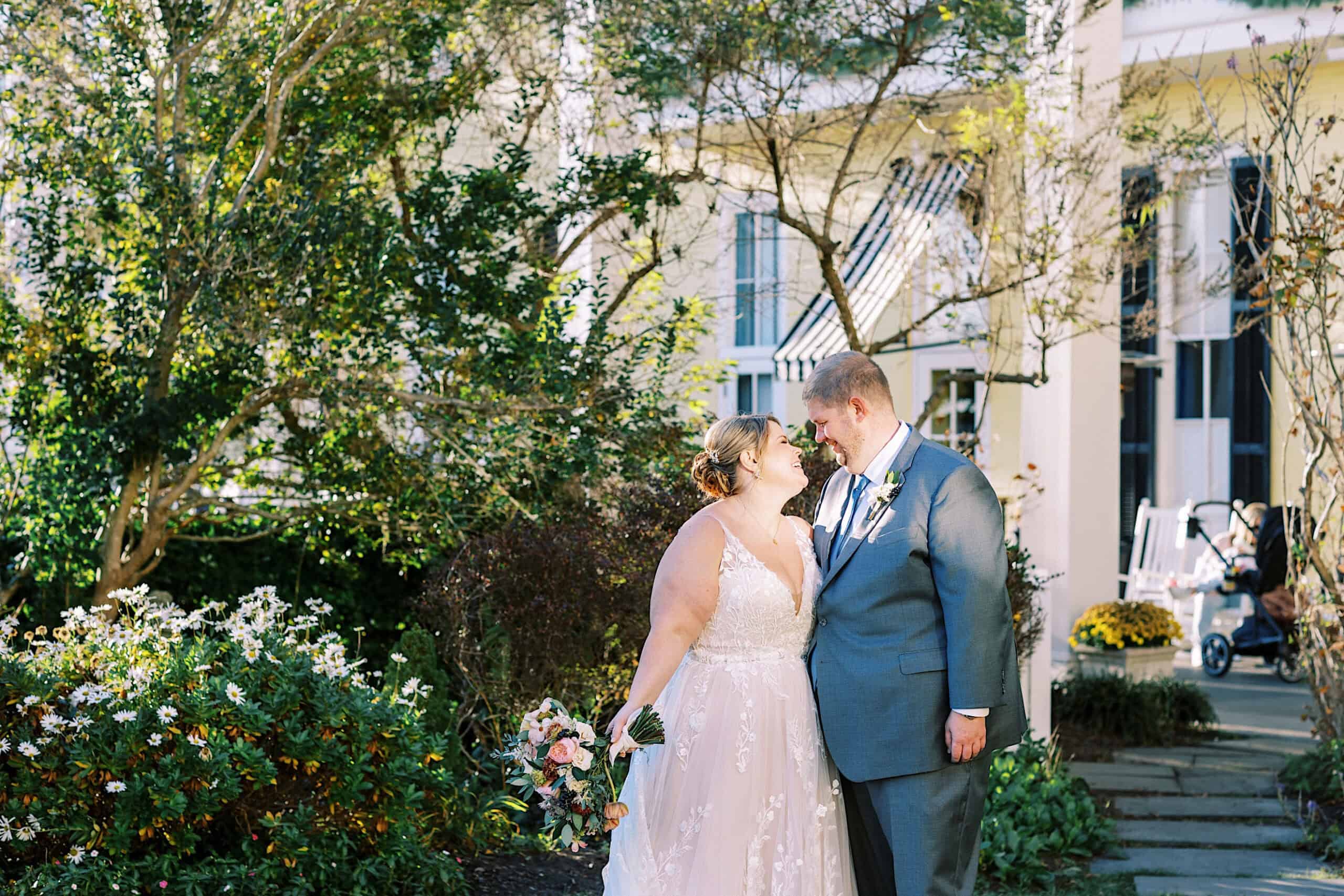 A bride and groom stand close together outdoors in a garden, smiling at each other, surrounded by flowers and greenery during their romantic November wedding at Congress Hall.