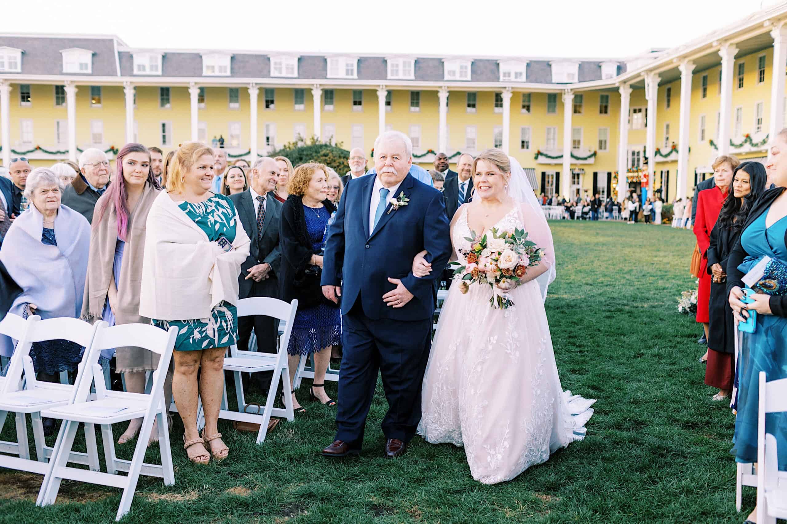 A bride in a white gown walks arm-in-arm with an older man down an outdoor aisle at Congress Hall, surrounded by guests, during a romantic November wedding in front of a large yellow building.