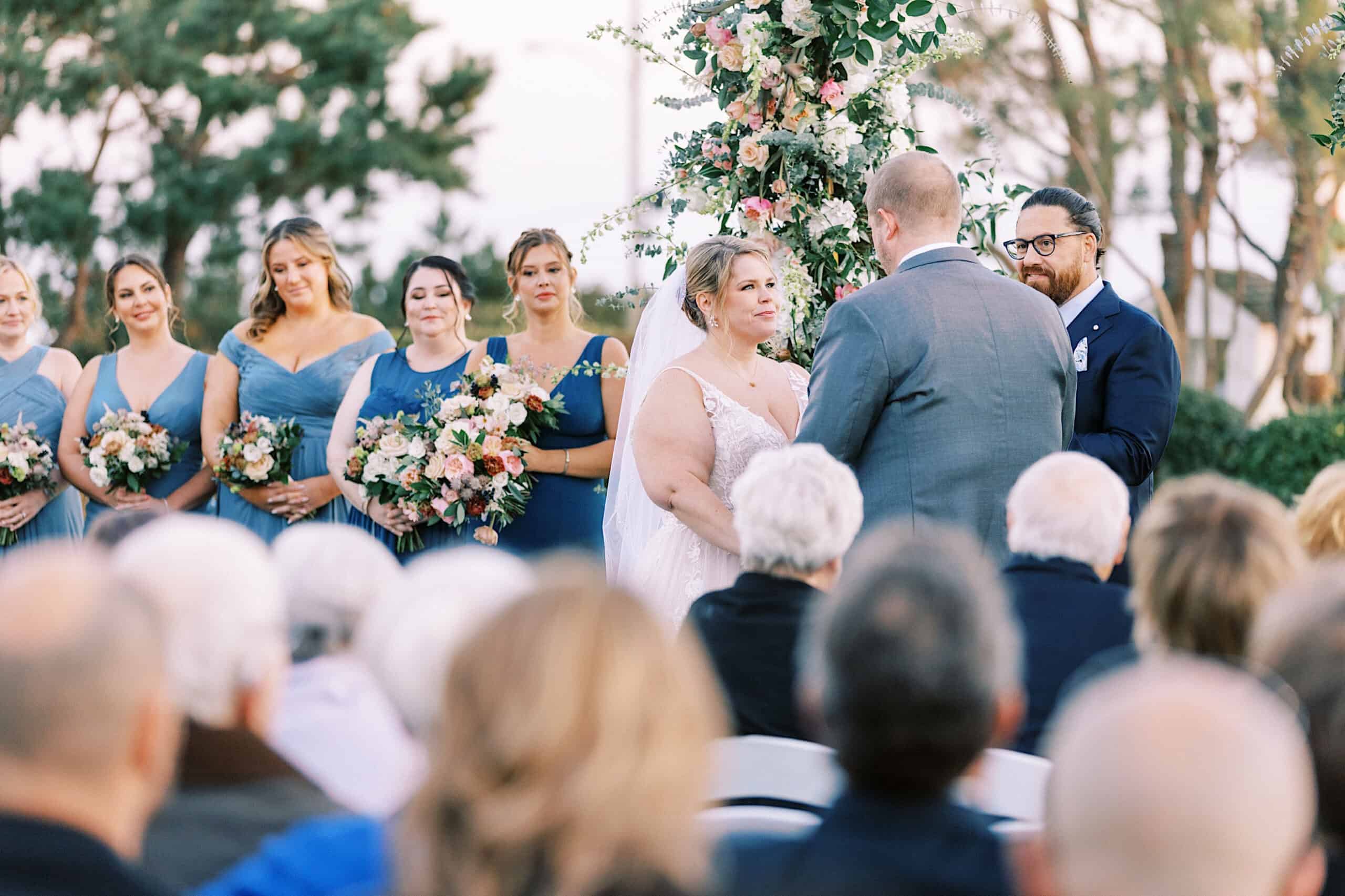 A bride and groom stand facing each other at a romantic November wedding at Congress Hall, surrounded by bridesmaids in blue dresses and guests seated in the foreground.