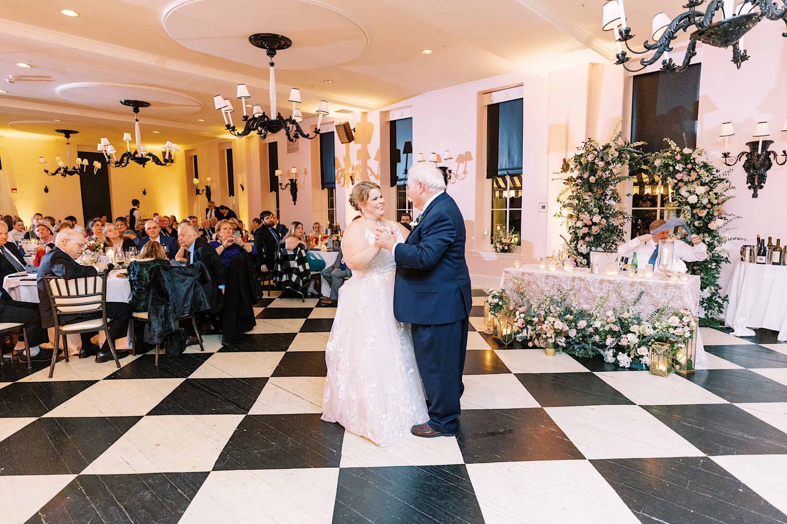 A bride and groom share a first dance on a black-and-white checkered floor in a decorated ballroom, as guests look on—a romantic November wedding at Congress Hall.