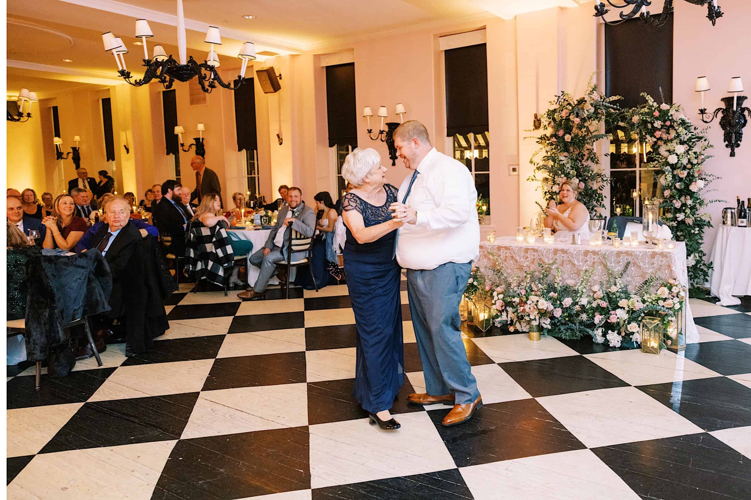 At a romantic November wedding at Congress Hall, a man and an elderly woman dance together on a black-and-white checkered floor as guests watch from elegantly decorated tables.