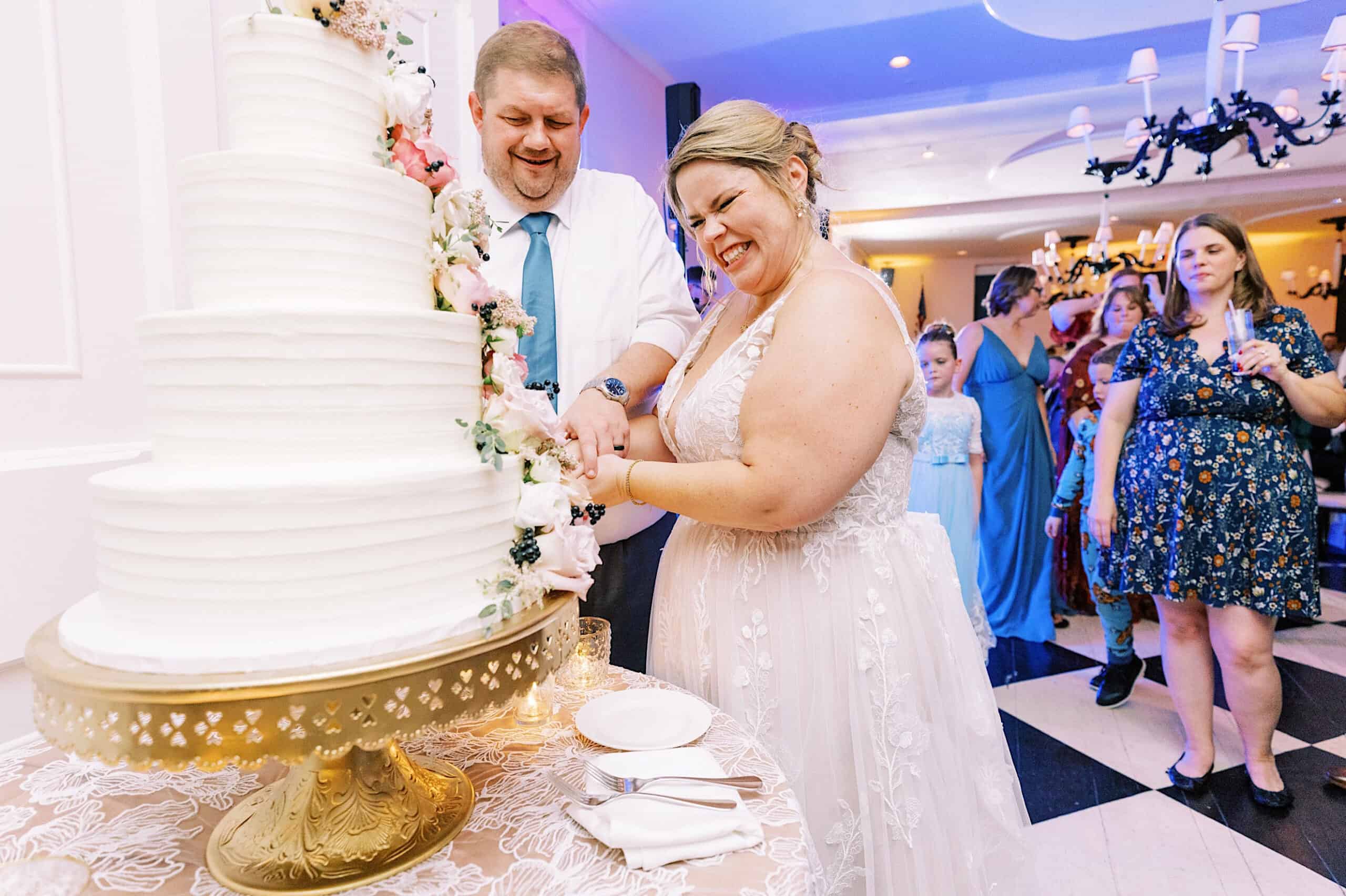 A bride and groom smile as they cut a tiered white wedding cake together during their romantic November wedding at Congress Hall, with guests watching in the background at the beautifully decorated indoor venue.