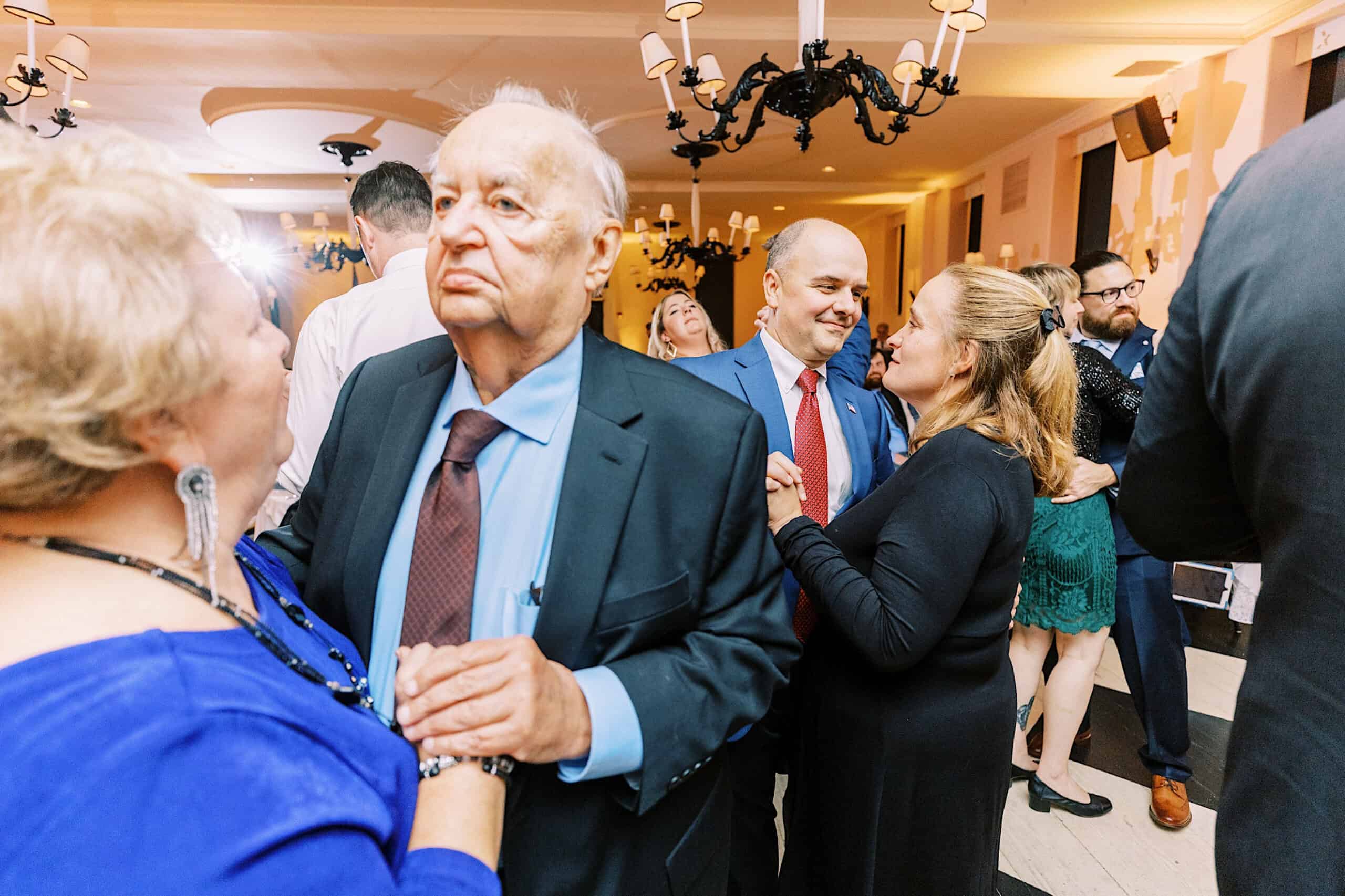 Several people are dancing in a well-lit indoor venue during a romantic November wedding at Congress Hall, dressed in formal attire, with chandeliers overhead and others mingling in the background.